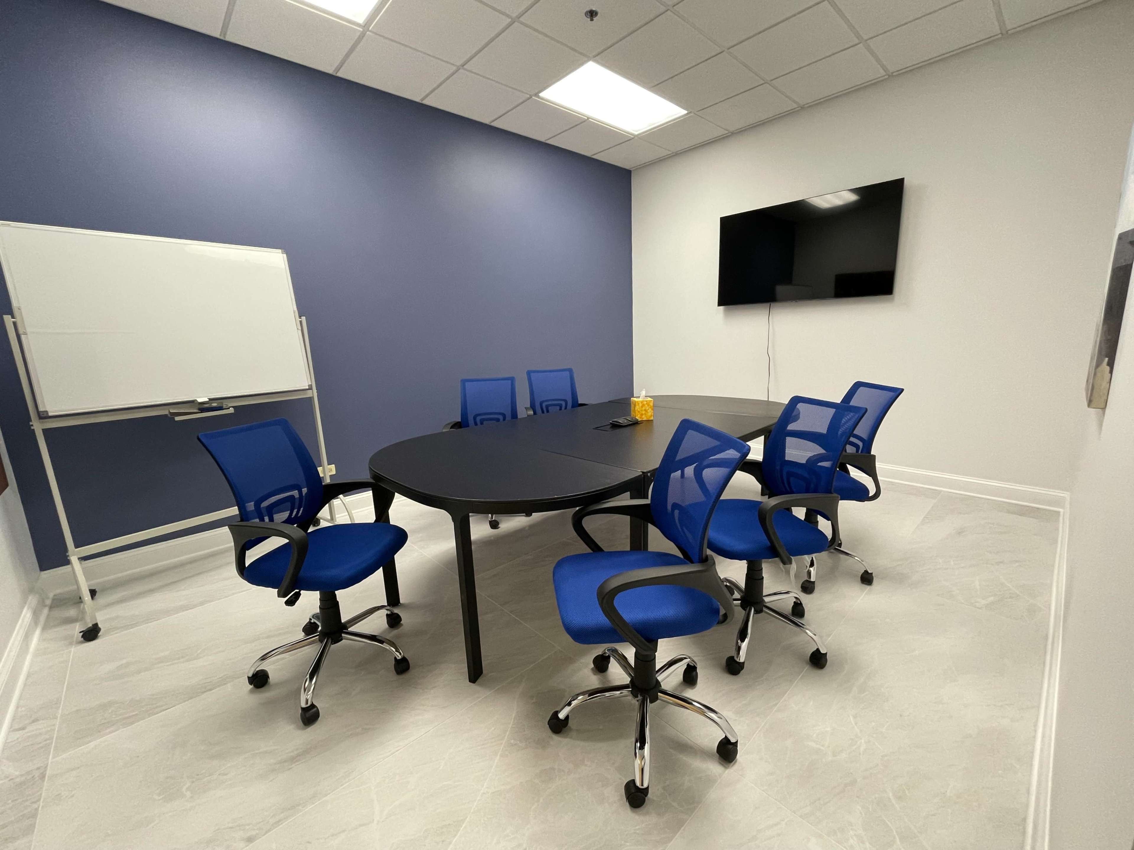 A modern conference room features a large oval table surrounded by six blue rolling chairs, a whiteboard, and a wall-mounted television.