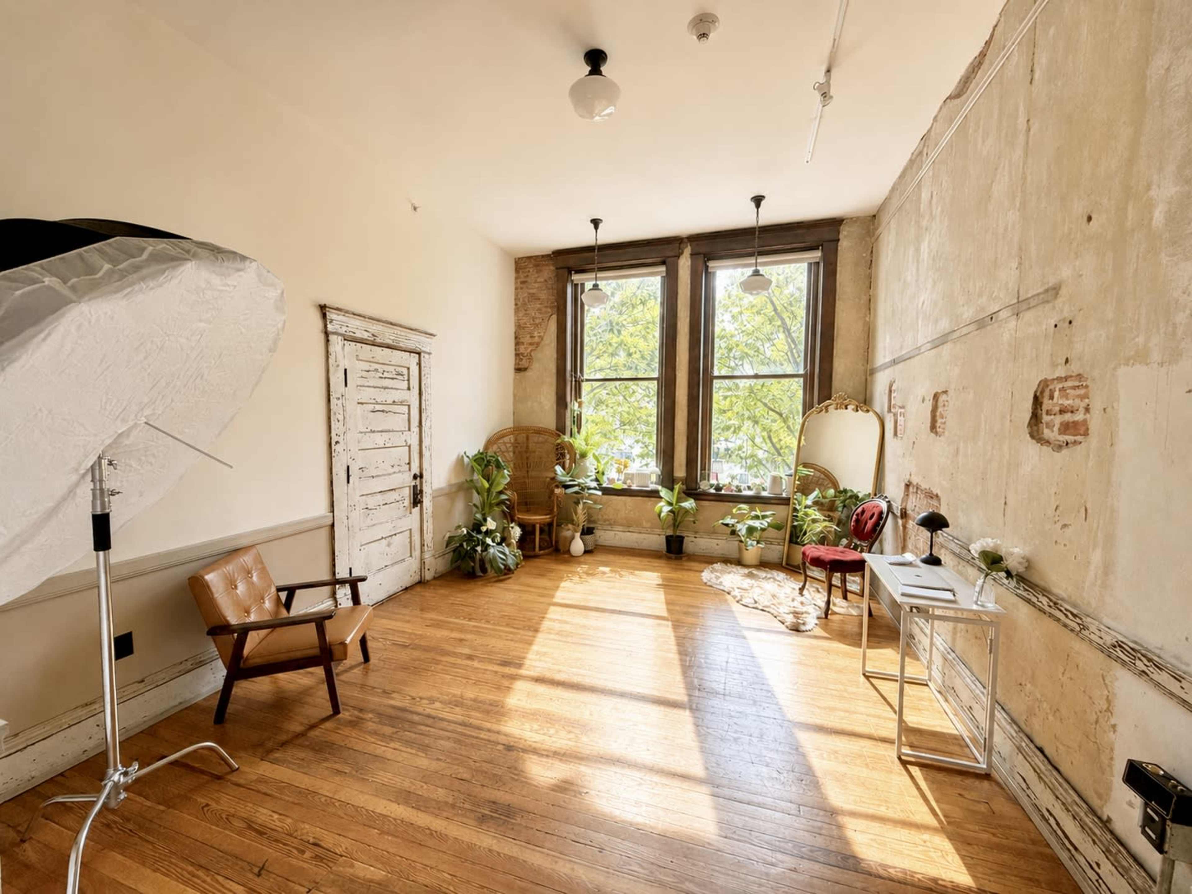 The image shows a bright, empty room with wooden floors, large windows, and minimal furniture, including a chair and a small table.