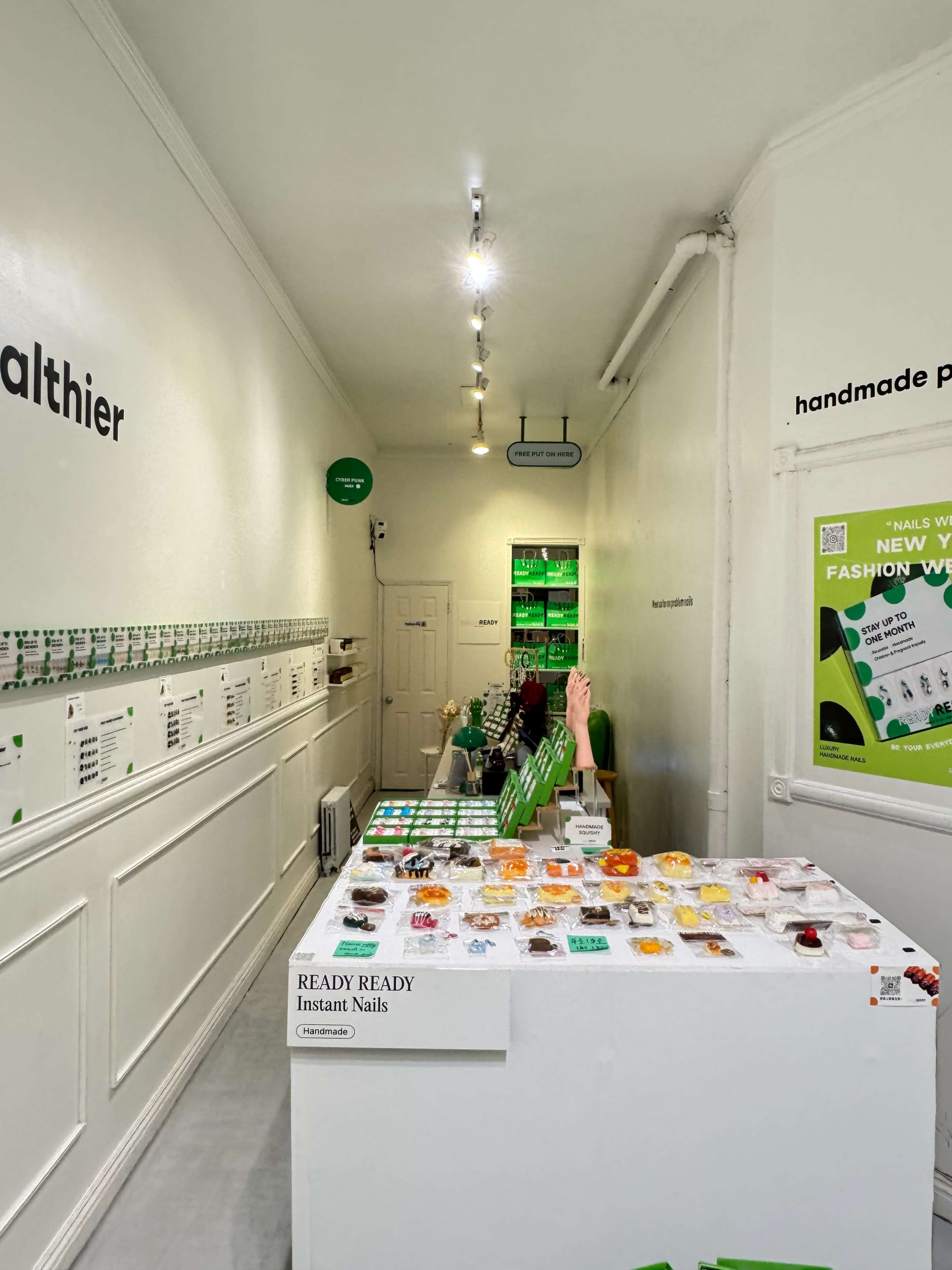 The image shows a brightly lit store interior with white walls, featuring a display table filled with colorful nail products and a selection of items arranged on the walls.