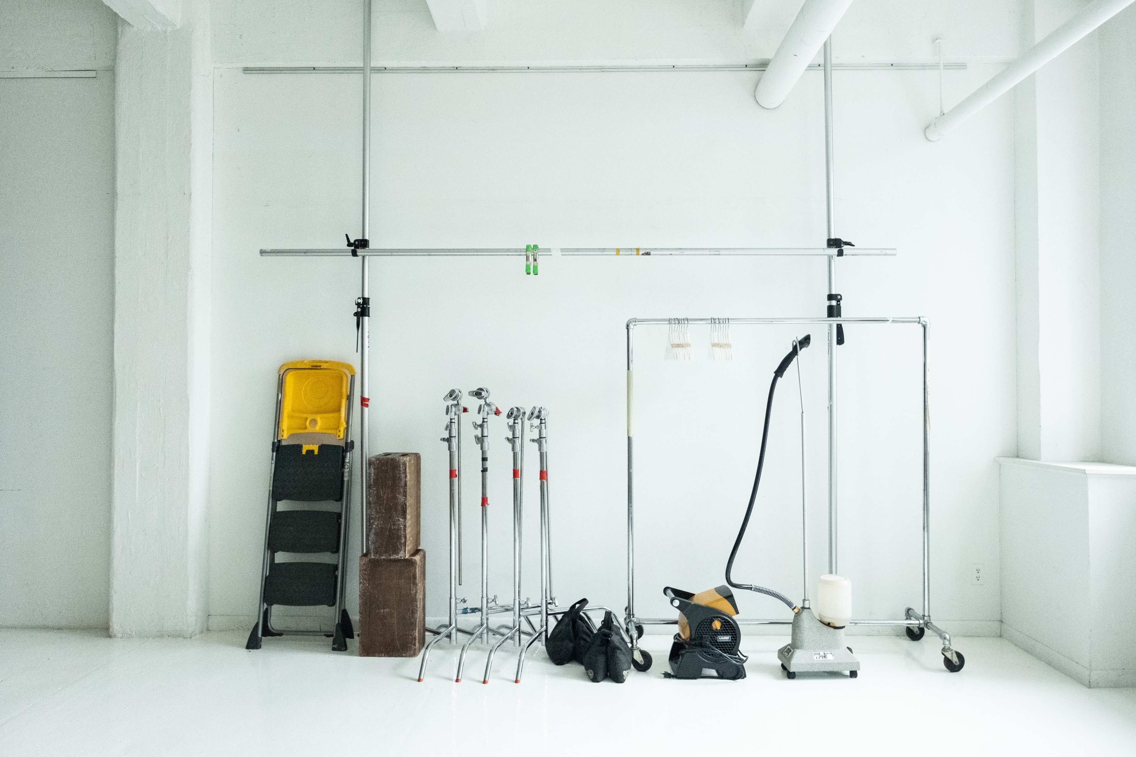 A collection of equipment, including light stands, a ladder, a vacuum cleaner, and a fabric backdrop support, is arranged against a white wall in a minimalist space.