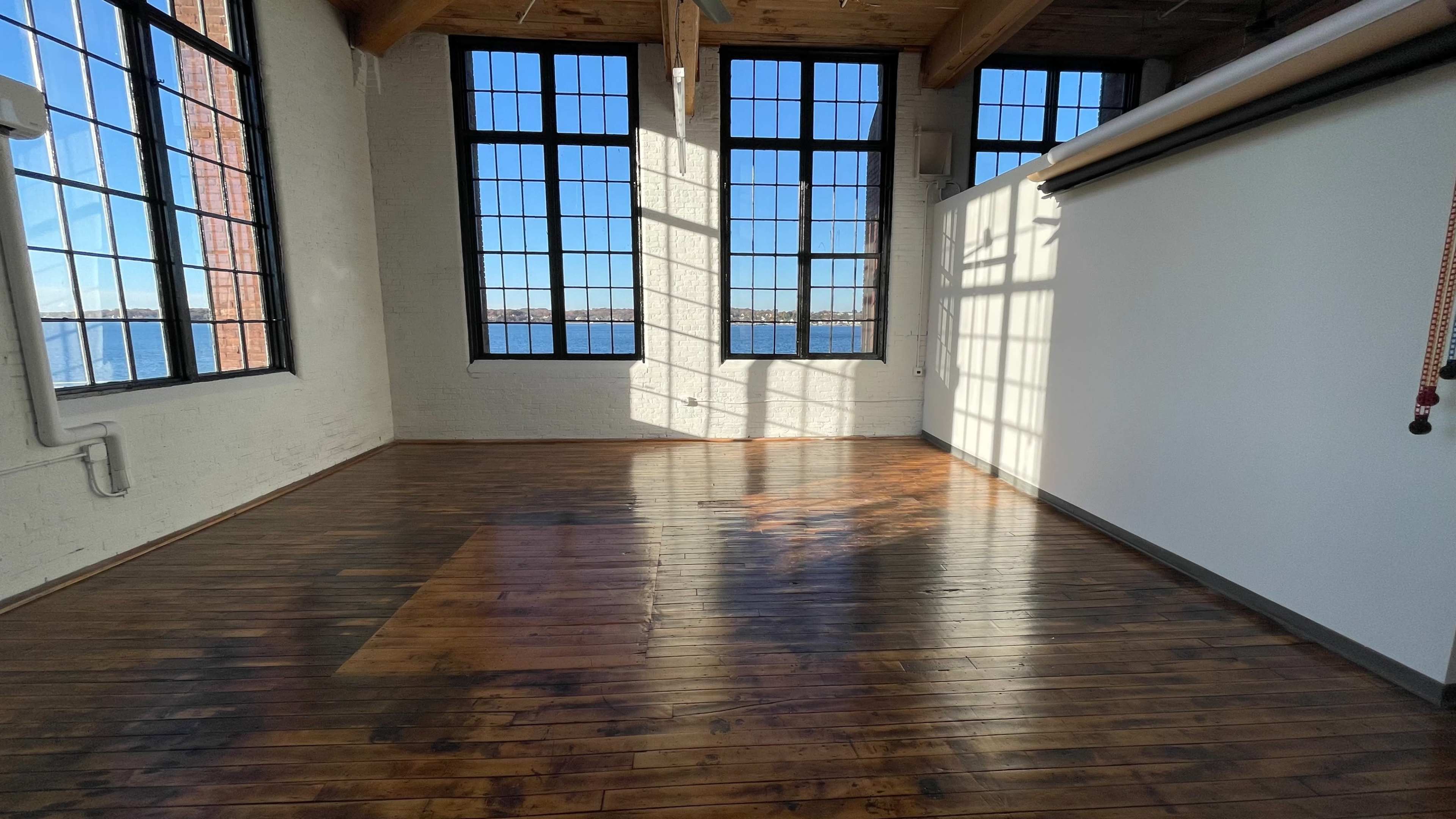 An empty room with large windows along one wall, showcasing wooden floors and a view of water outside.