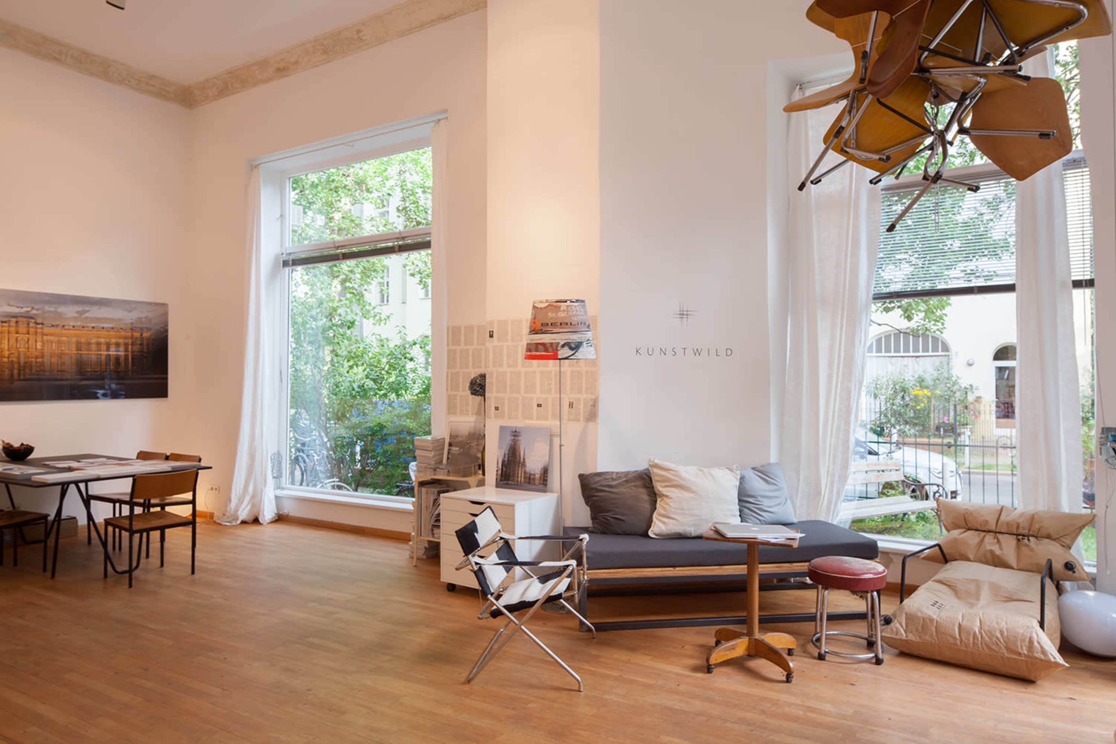 A spacious, bright room featuring a mix of modern furniture, large windows with greenery outside, and a collection of art on the walls.