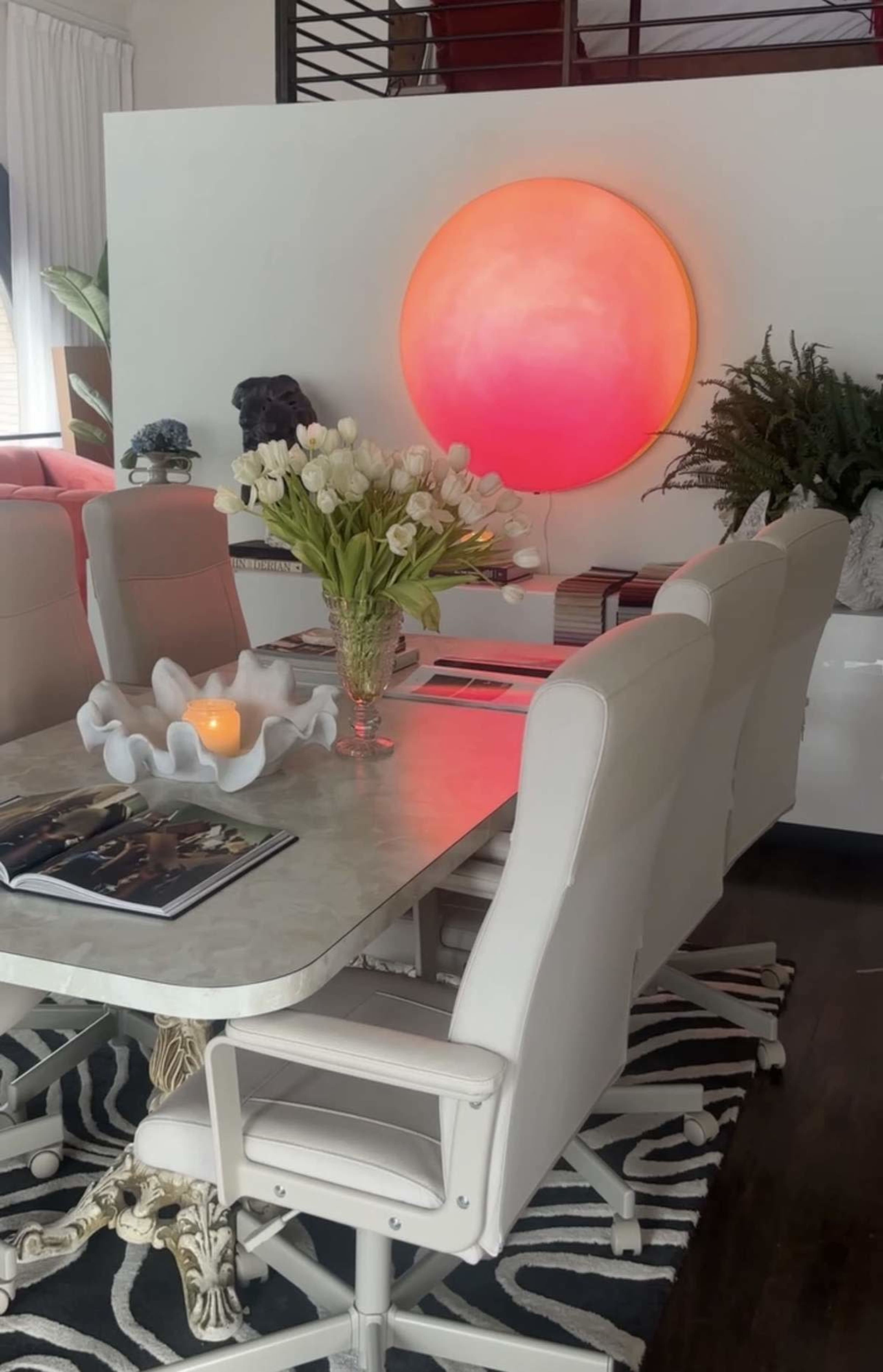 A dining table with white chairs is set in a room featuring a large circular red light on the wall and a vase of white flowers in the center.