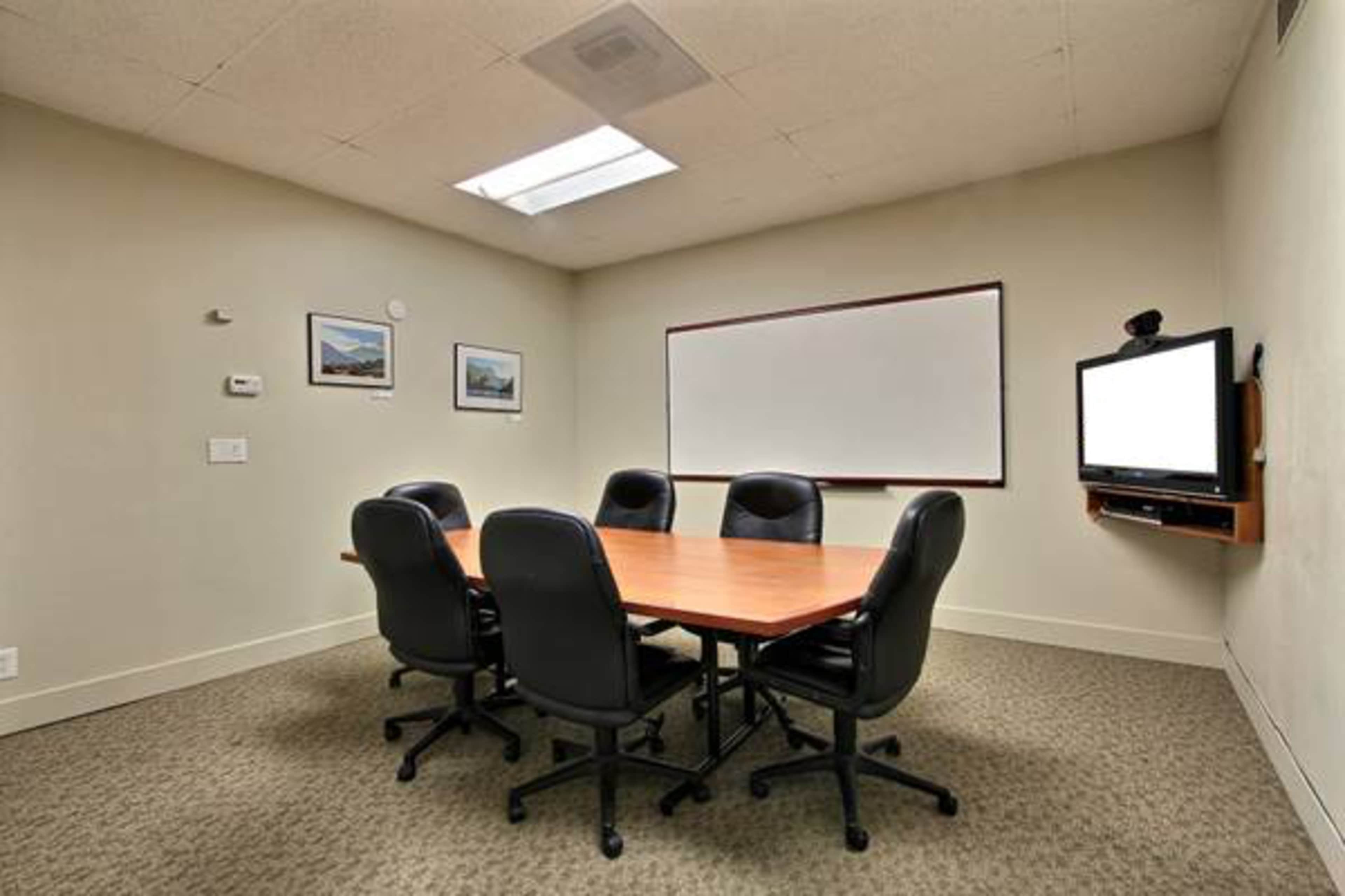 A small conference room features a rectangular wooden table surrounded by six black chairs, with a whiteboard and a television mounted on the wall.