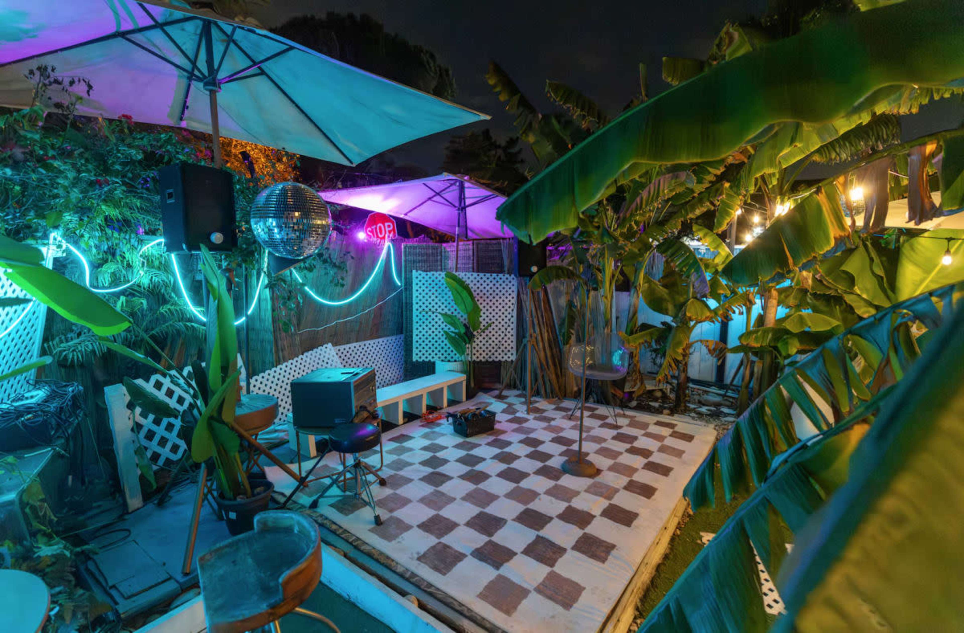 The image shows an outdoor party space with checkered flooring, colorful neon lights, a disco ball, and tropical plants.