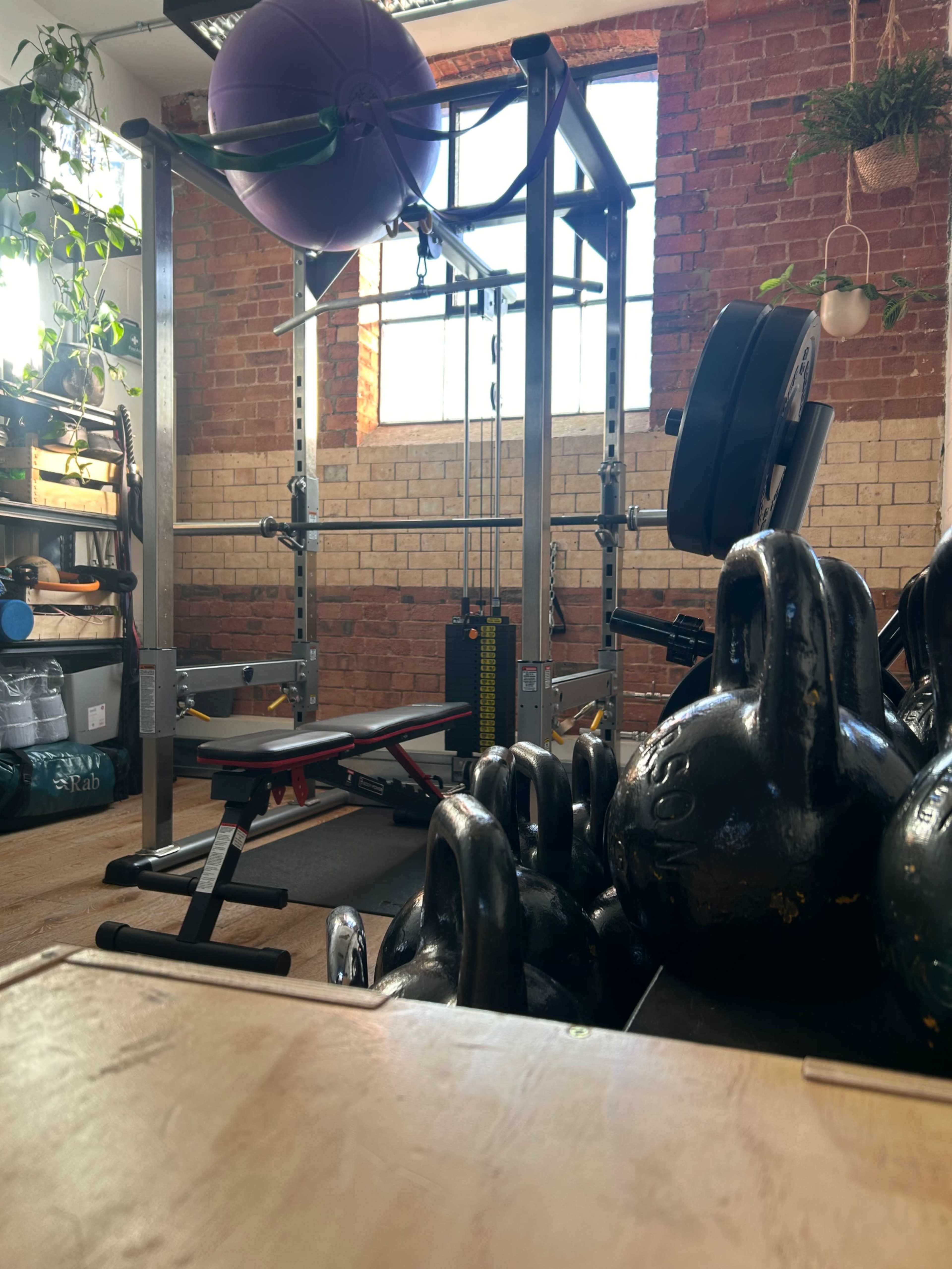 The image shows a well-equipped gym area featuring a weightlifting rack, a bench, kettlebells, and a large exercise ball against a brick wall with windows.