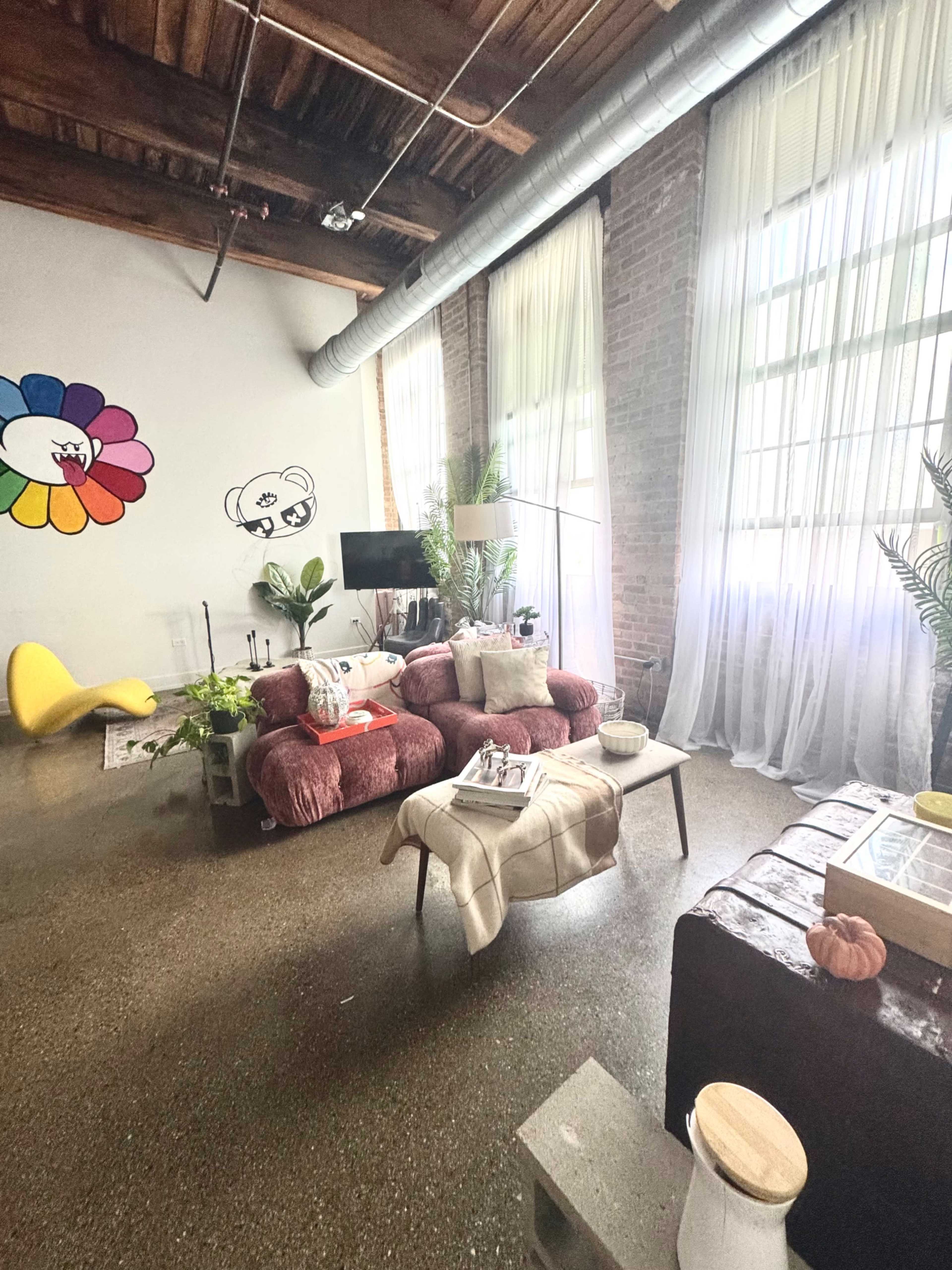 The image shows a bright living room with a red sofa, a coffee table, and large windows draped with sheer curtains, complemented by plants and artwork on the walls.