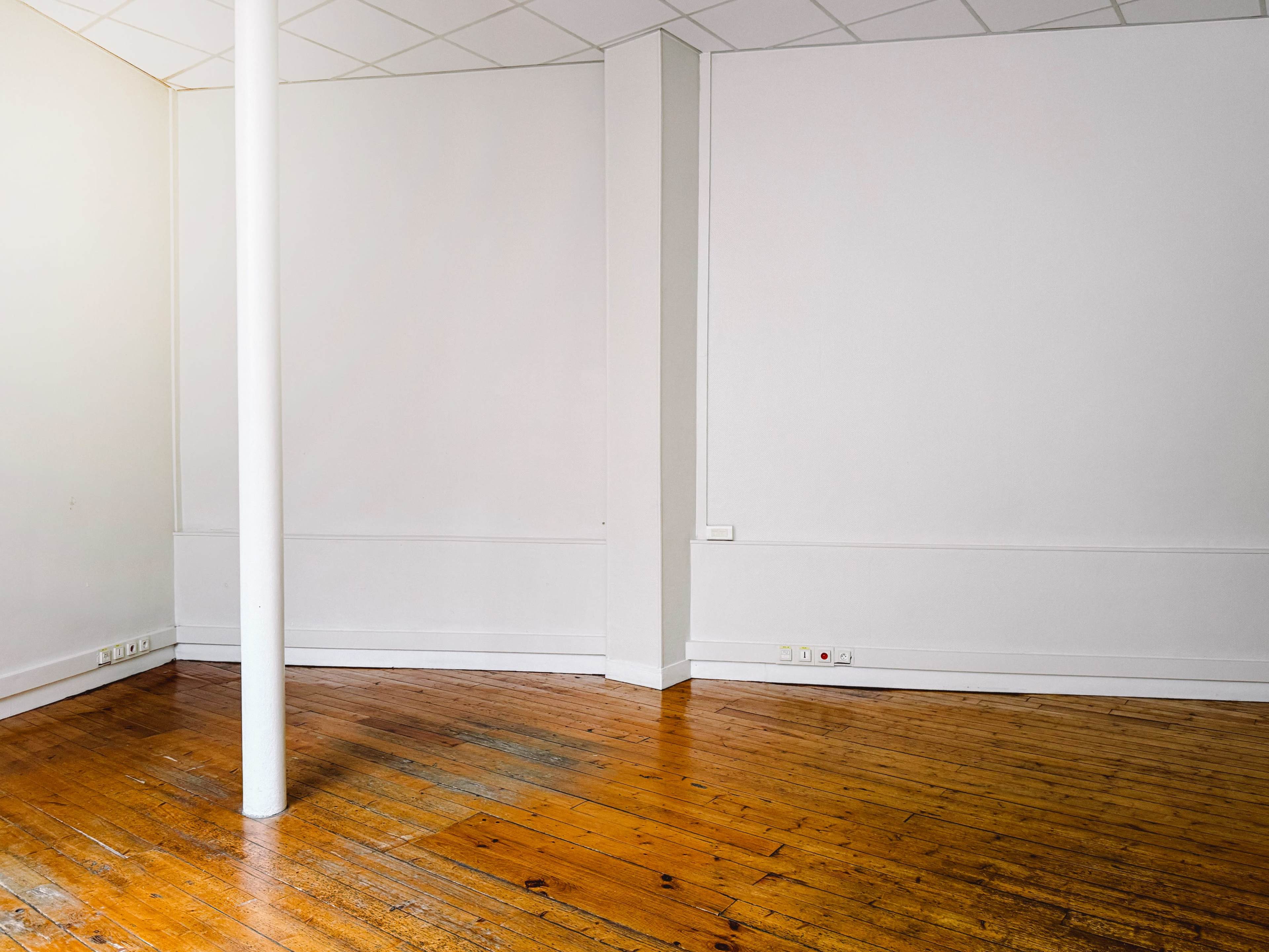 The image shows an empty room with wooden floors, a white column, and plain, light-colored walls.