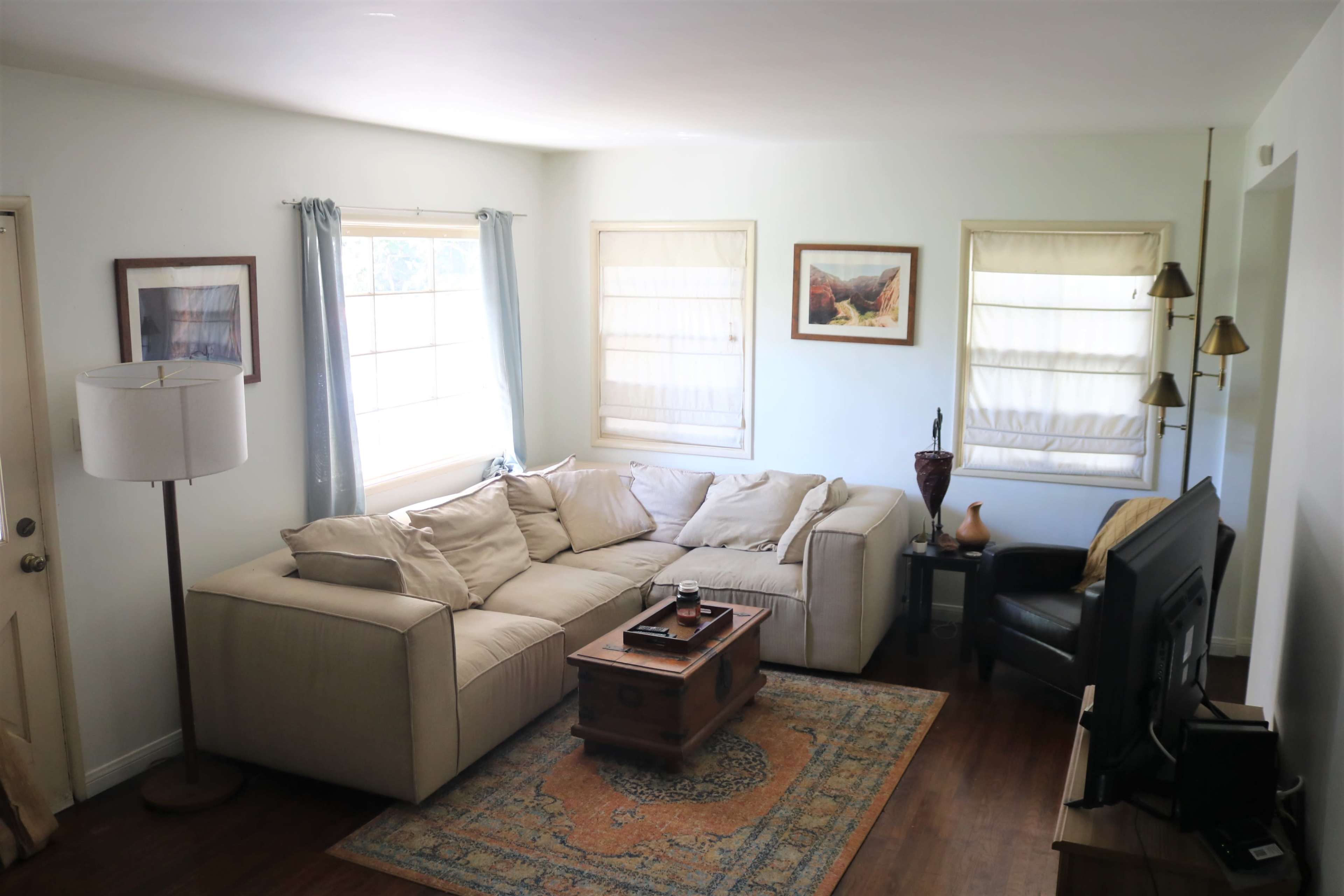 A cozy living room featuring a sectional sofa, a wooden coffee table, and a TV against the wall, with large windows allowing natural light to enter.