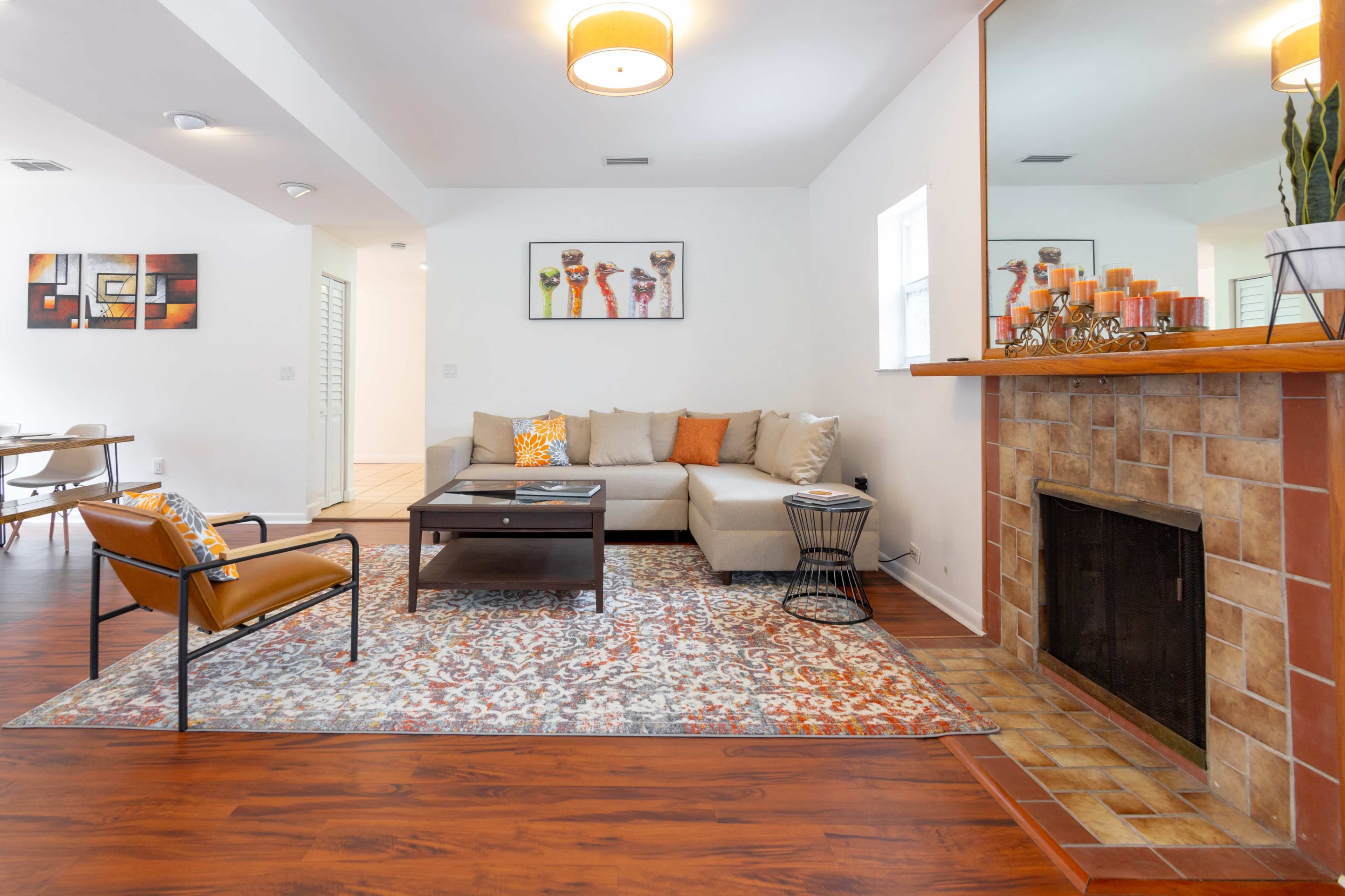 A living room with a beige sofa, a wooden coffee table, decorative items on a fireplace, and a patterned area rug on a hardwood floor.