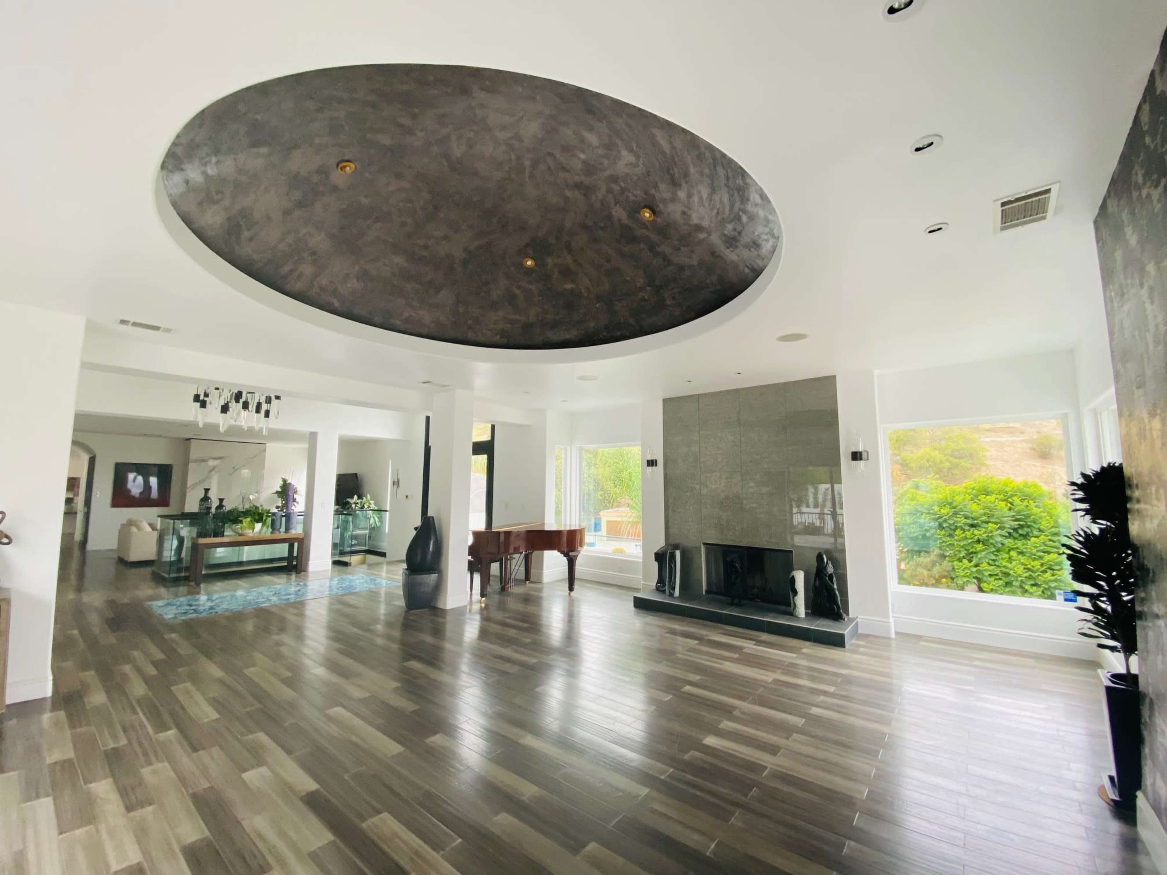 The image shows a spacious modern living area with a circular ceiling design, a fireplace, and large windows letting in natural light.