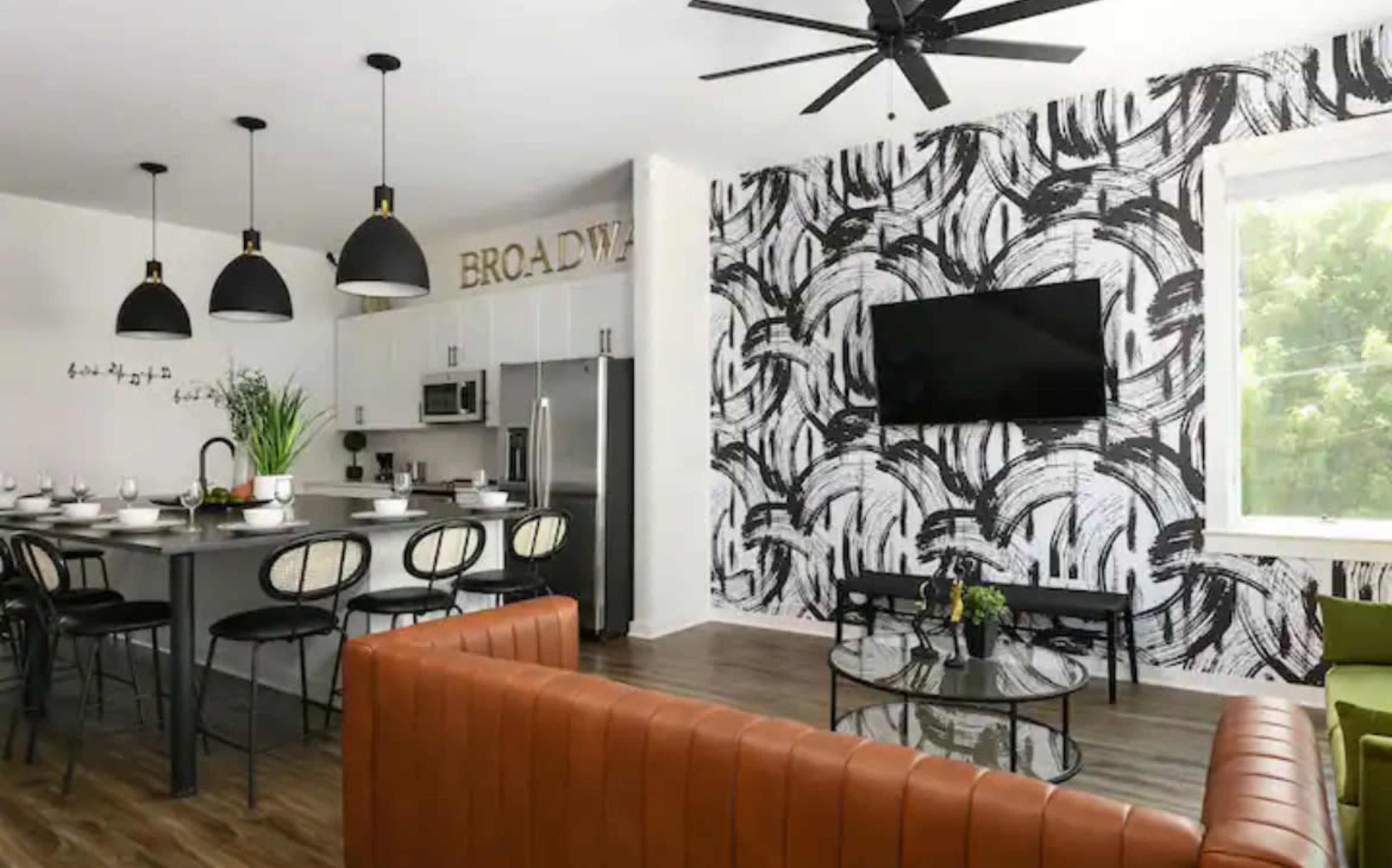 A modern open-concept living area featuring a kitchen island with bar seating, a dining table set for several guests, and a lounge area with a brown leather sofa and a coffee table, all against a patterned accent wall and a flat-screen TV.