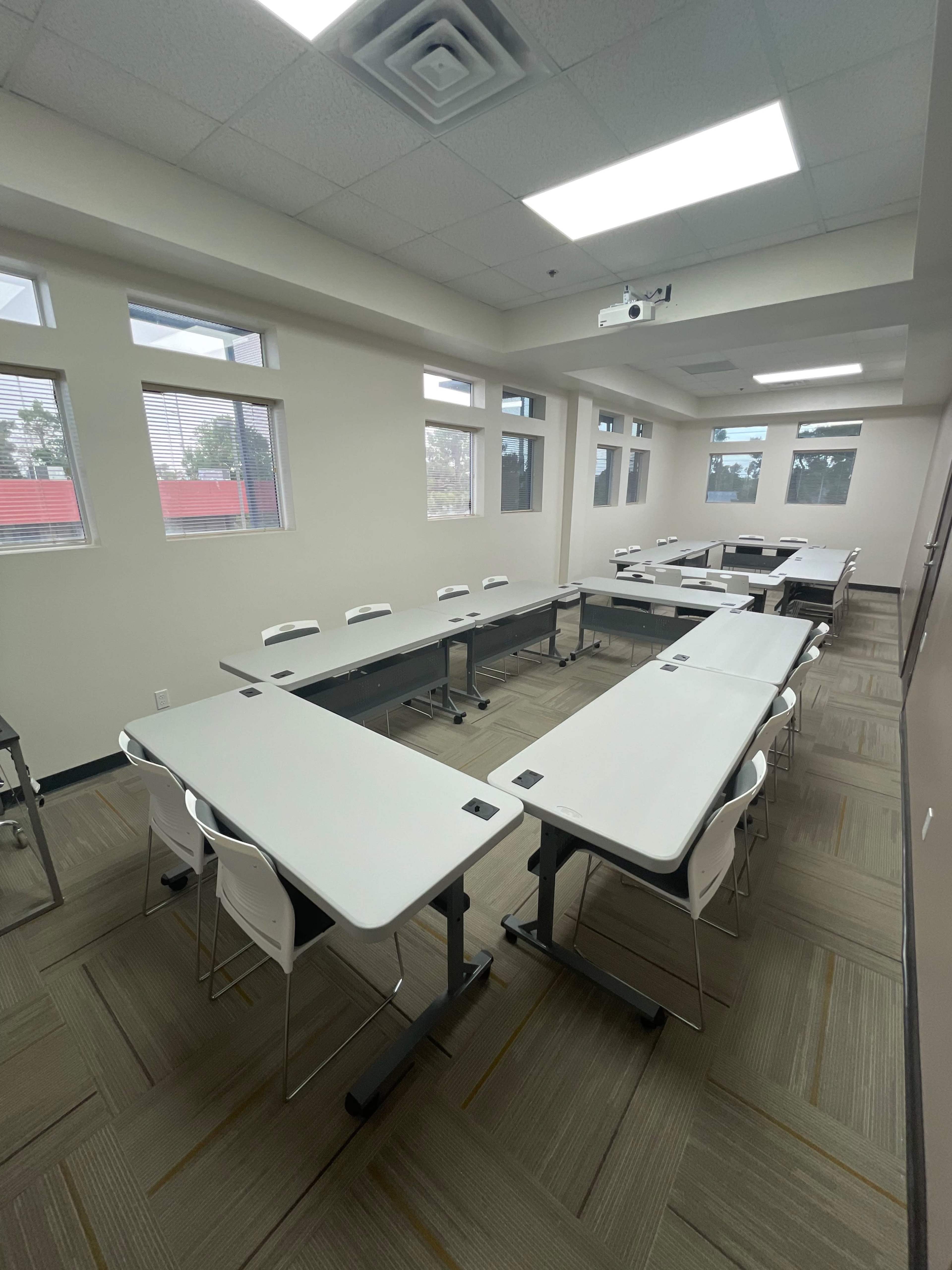 A classroom is arranged with multiple tables and chairs, featuring a projector on the ceiling and large windows for natural light.