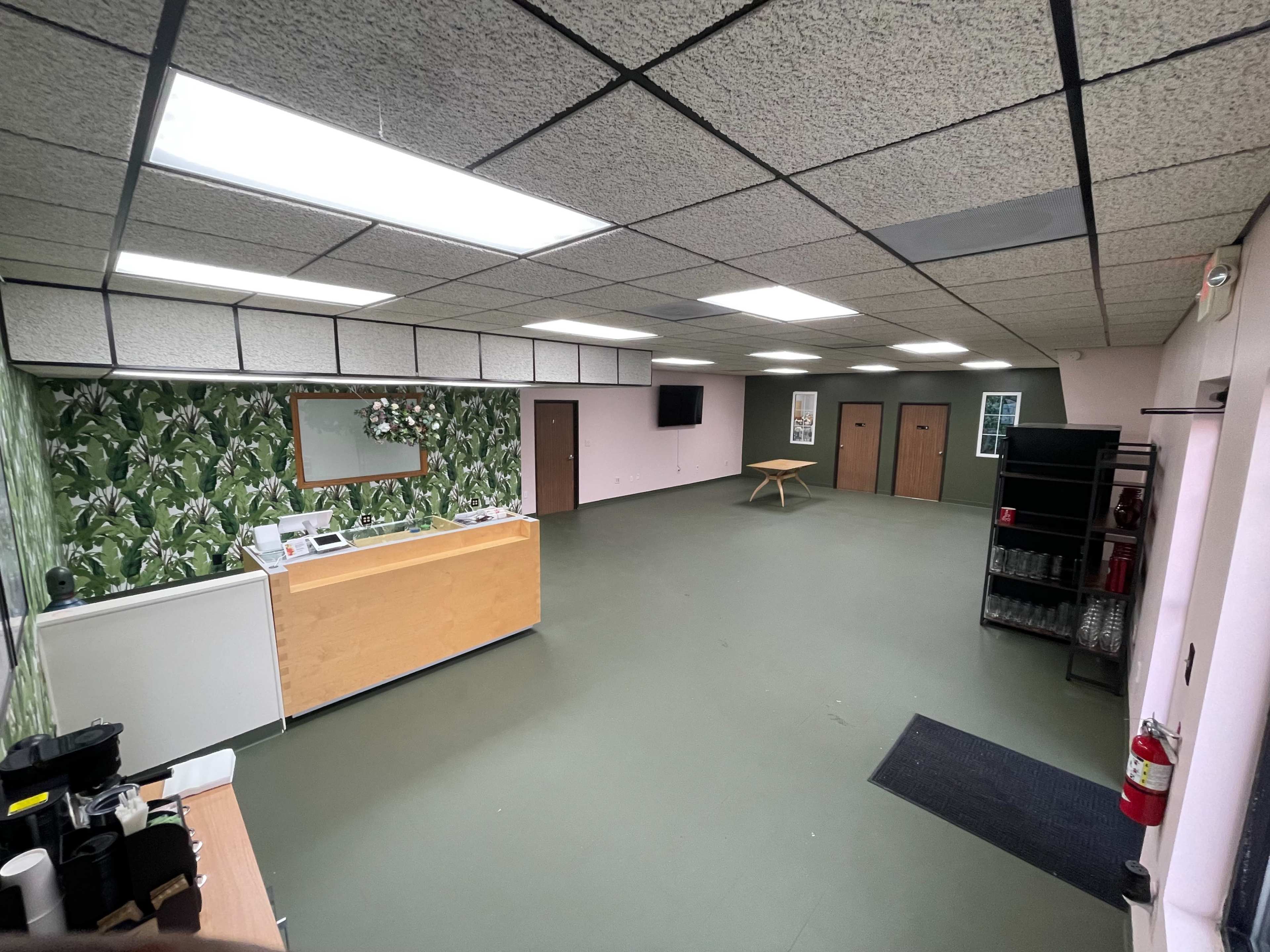 A spacious room with green flooring, a reception area with a counter, a wall decorated with plant-patterned wallpaper, and a central table surrounded by empty space.