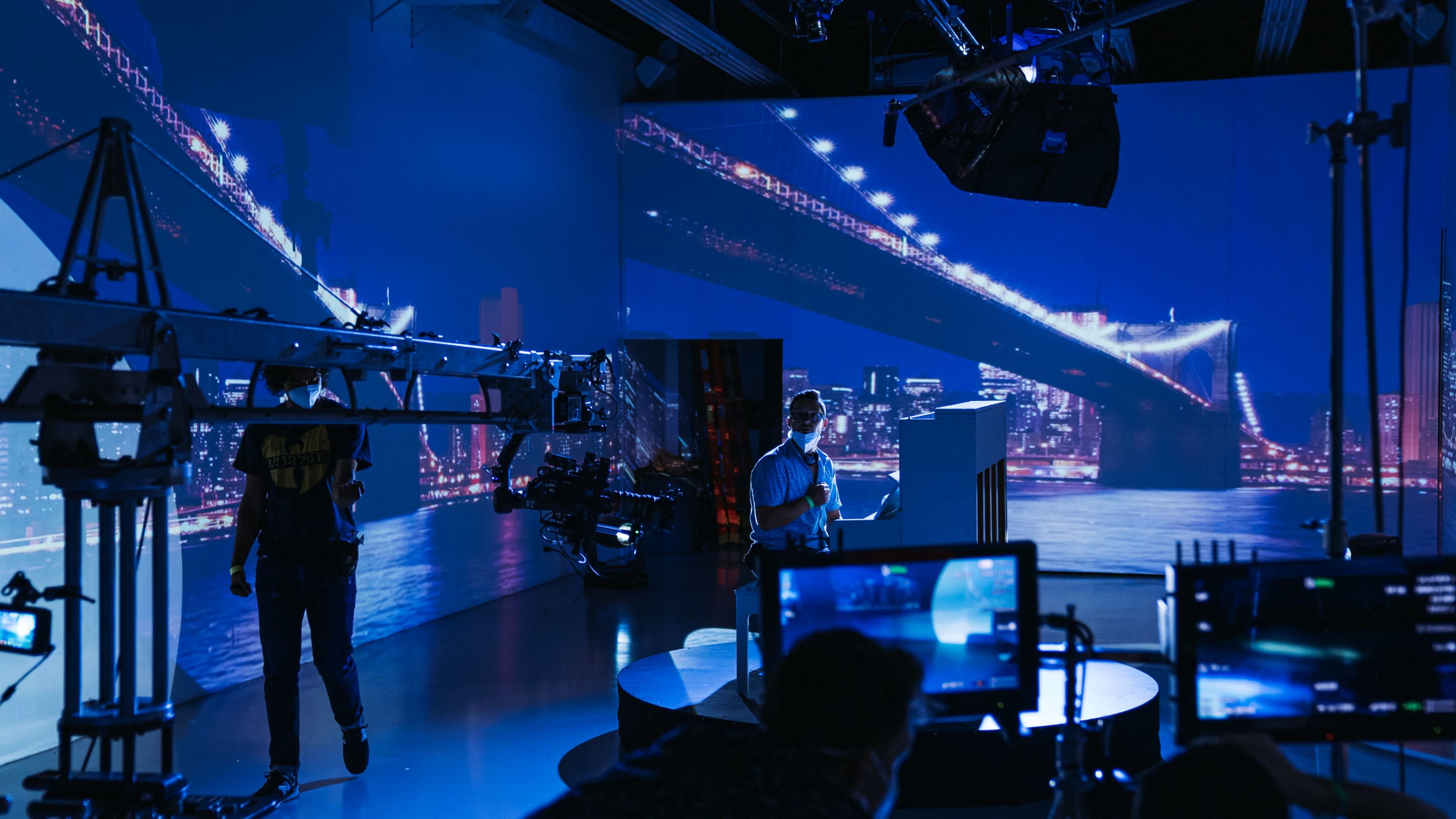 A television studio setup features a presenter on a circular podium with a backdrop of a brightly lit city skyline at night.