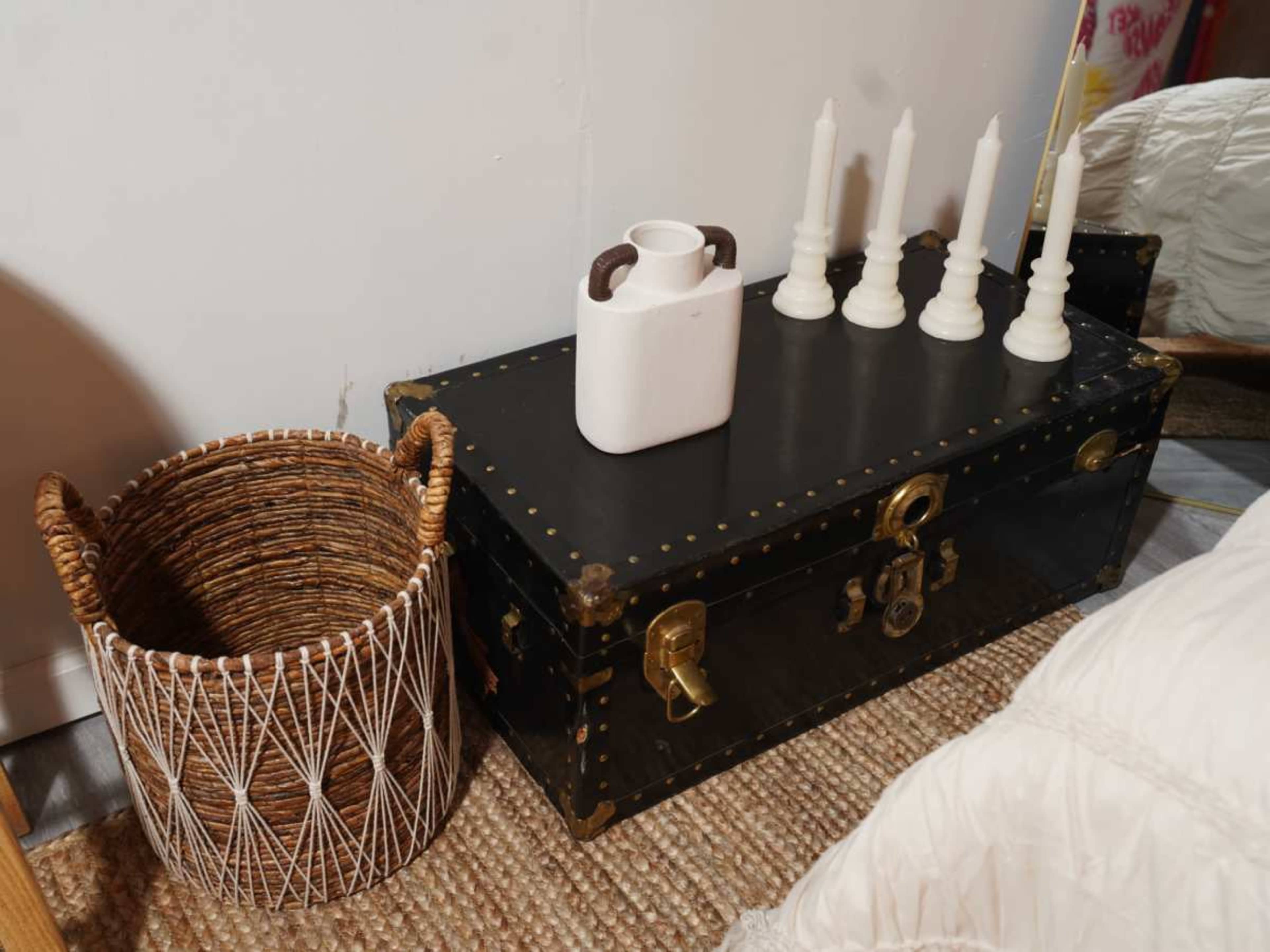 The image shows a black wooden trunk with brass accents, accompanied by a woven basket and four white candles in holders on top.