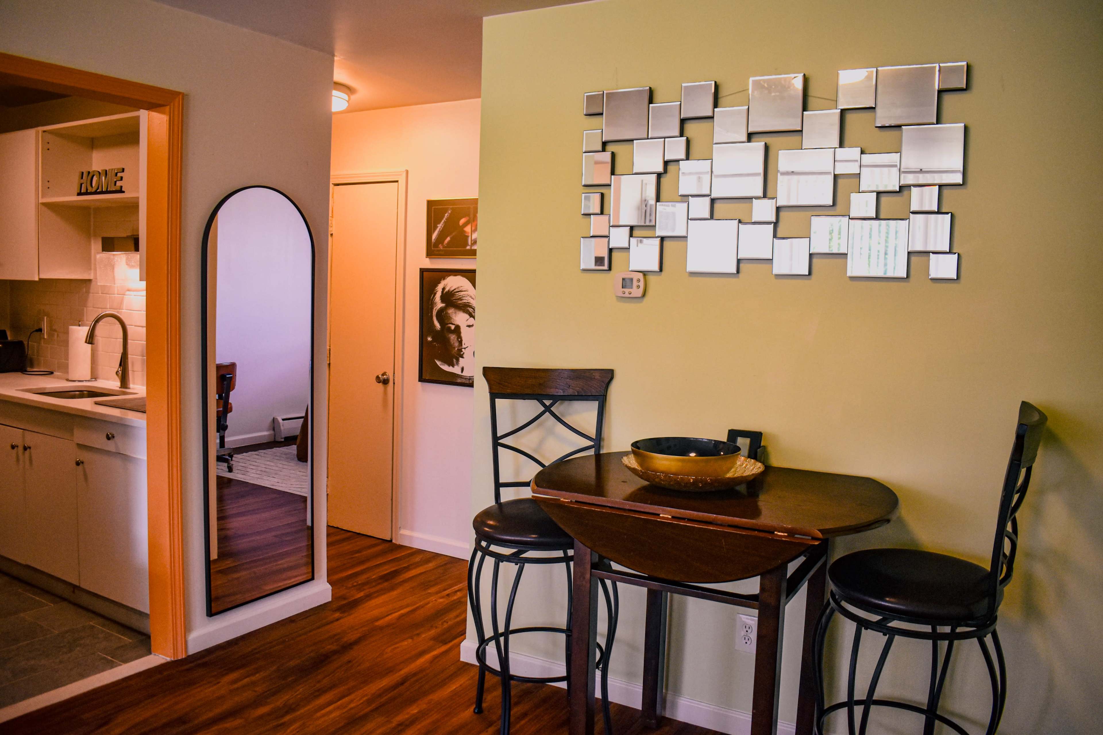 The image shows a compact dining area with a round table and two high-backed chairs, adjacent to a kitchen and a mirrored wall decoration.