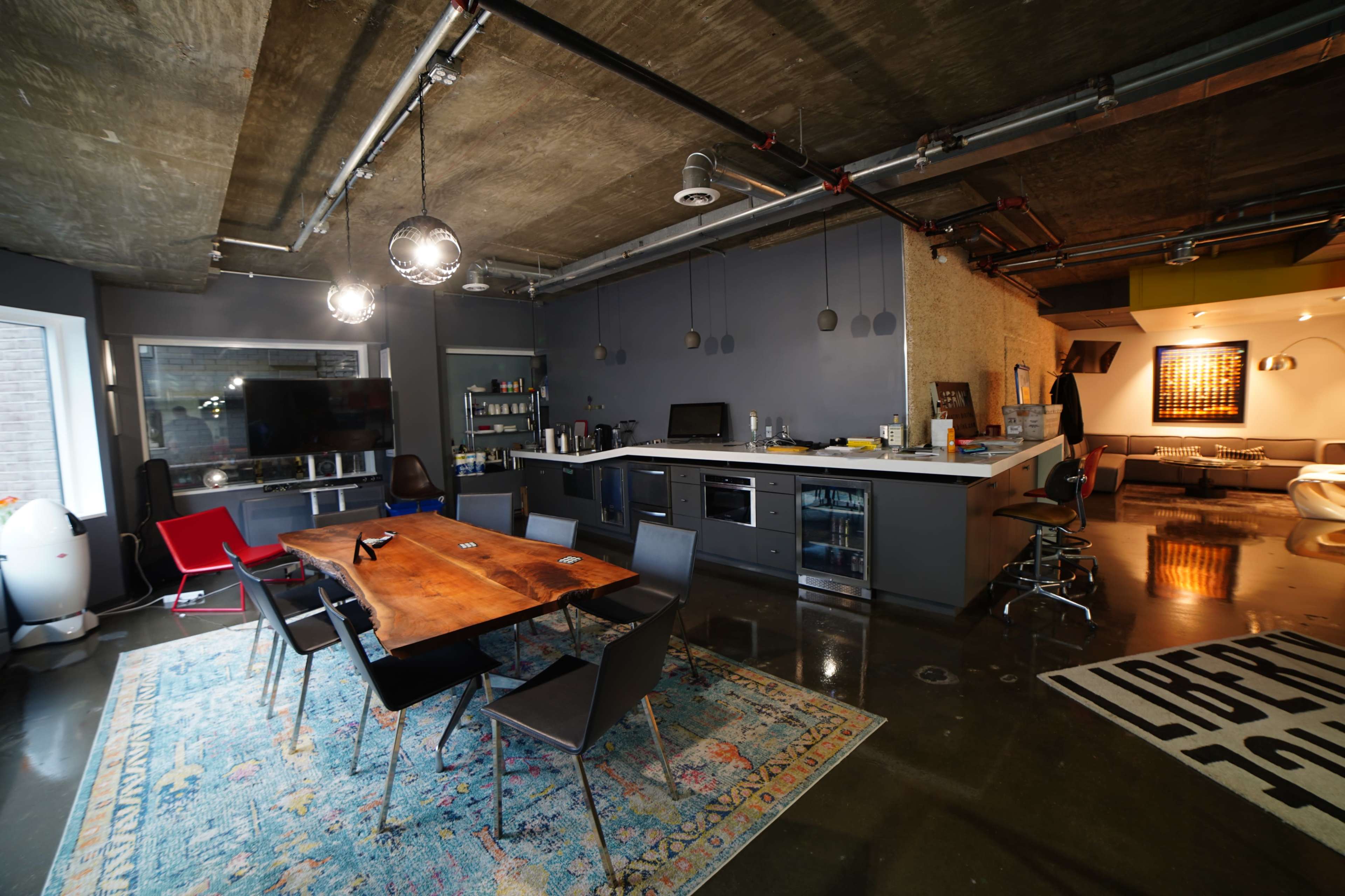 A modern, open-concept kitchen and dining area with a wooden table, black chairs, and sleek appliances, set against a backdrop of concrete walls and polished floors.