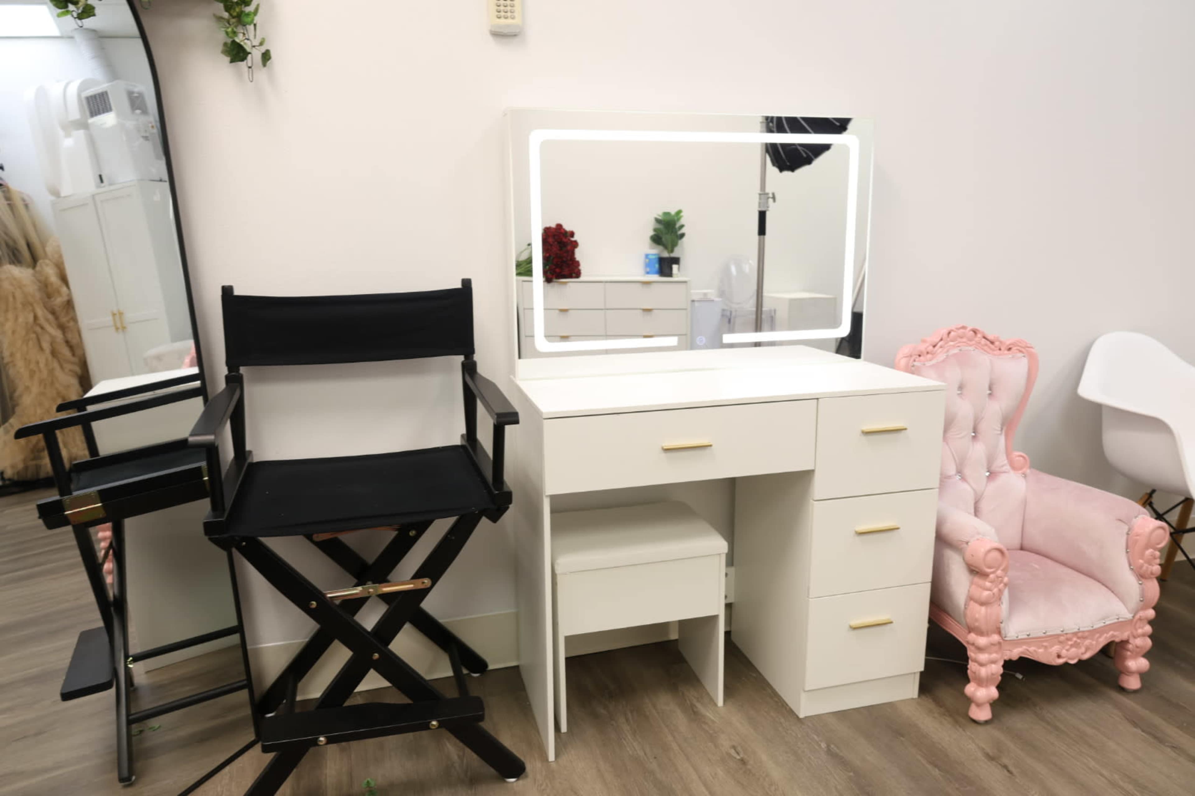 The image shows a makeup or dressing area featuring a black director's chair, a white vanity with a mirror, and a pink upholstered chair.