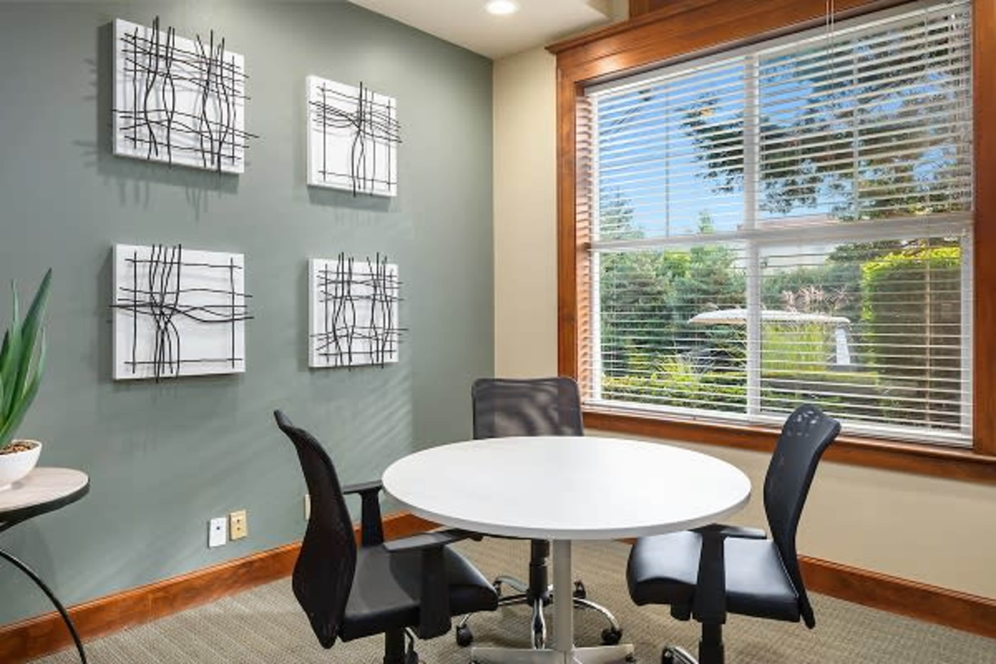 A round white table surrounded by two black chairs is positioned beside a window with blinds, offering a view of greenery outside, while three abstract wall art pieces are displayed on the green wall.