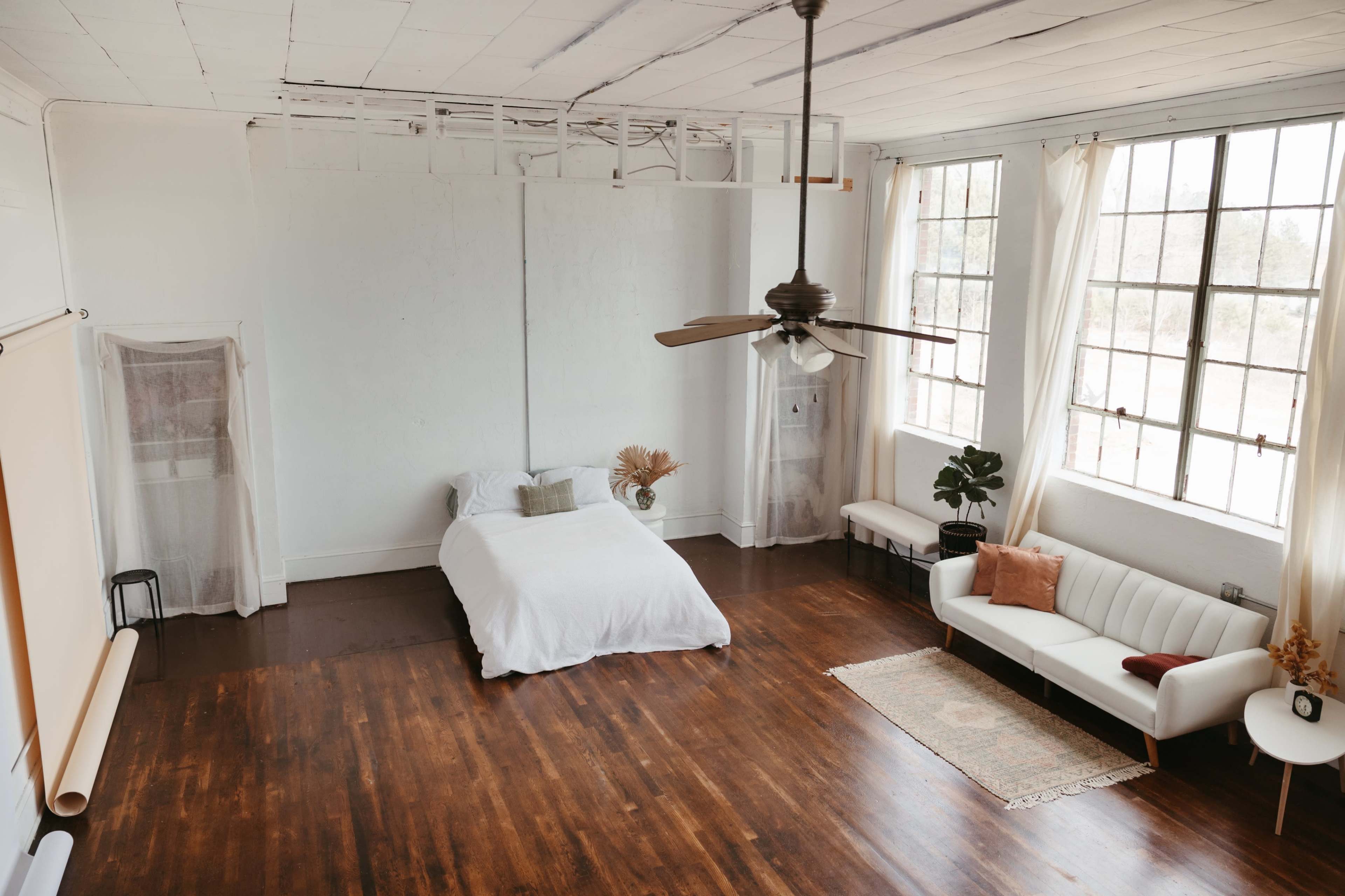 The image shows a spacious, minimally furnished bedroom with a bed, a sofa, and large windows allowing natural light to enter.