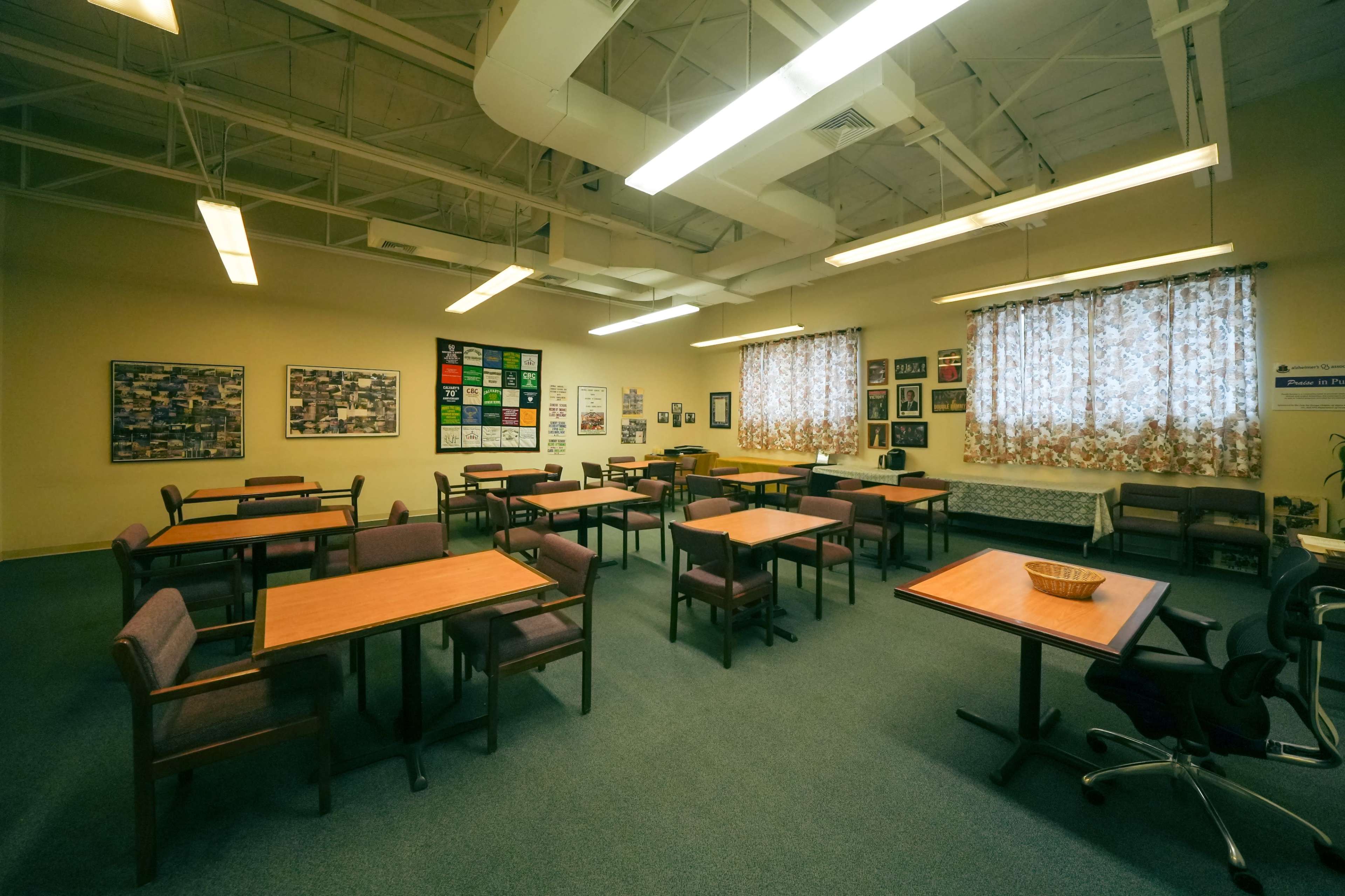 A spacious classroom with several wooden tables and chairs arranged throughout, large windows with patterned curtains, and various educational posters on the walls.