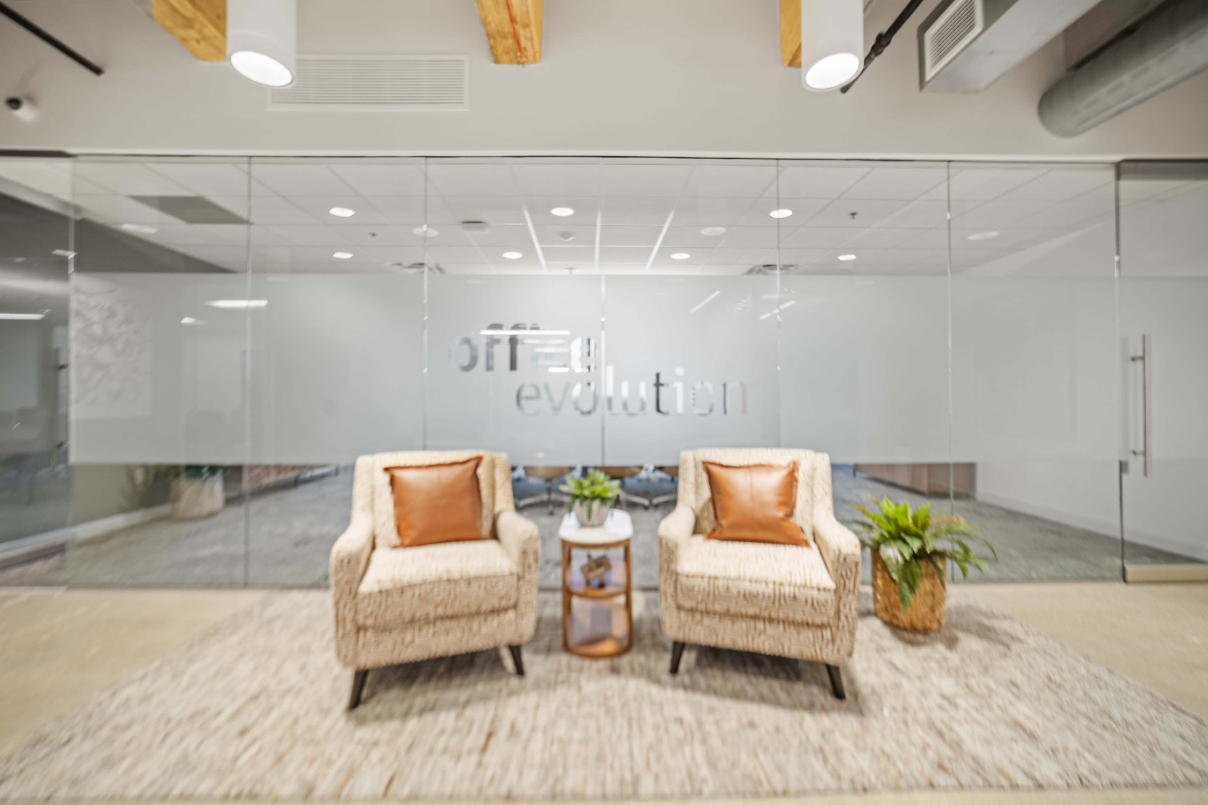 The image shows a modern office waiting area with two cream-colored armchairs and a small table surrounded by indoor plants.