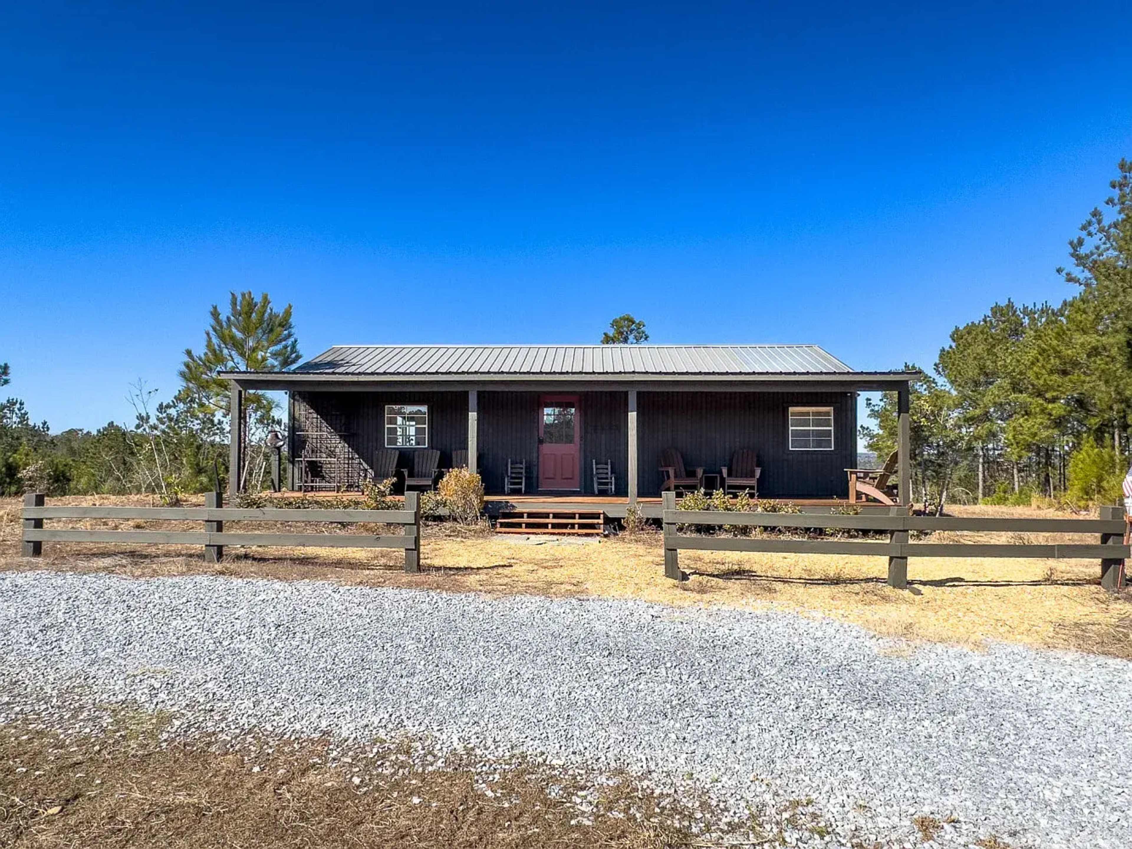 A black wooden house with a metal roof is set on a gravel driveway, surrounded by trees and open land.