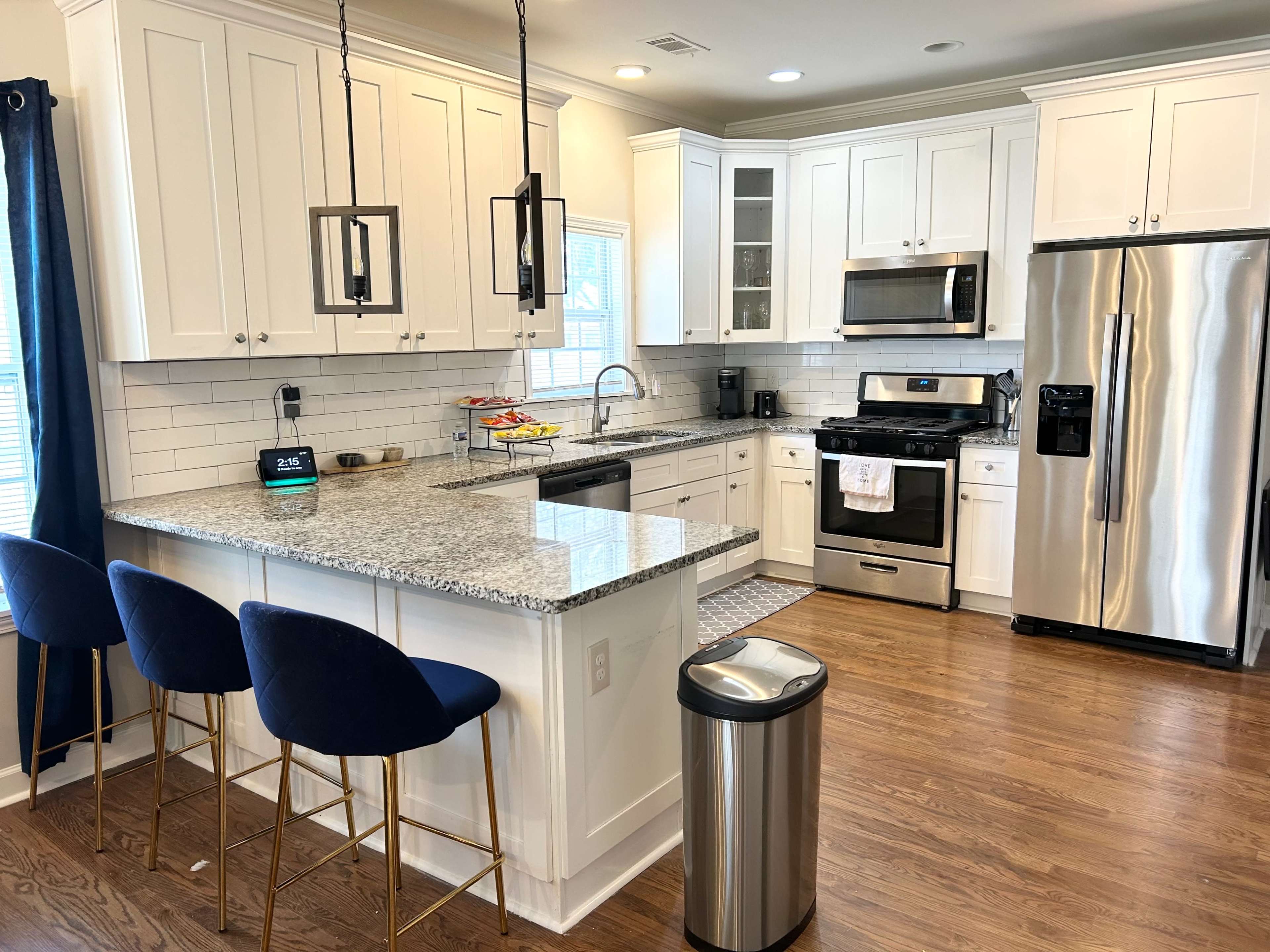 The image shows a modern kitchen with white cabinetry, stainless steel appliances, a granite countertop, and a central island with blue bar stools.