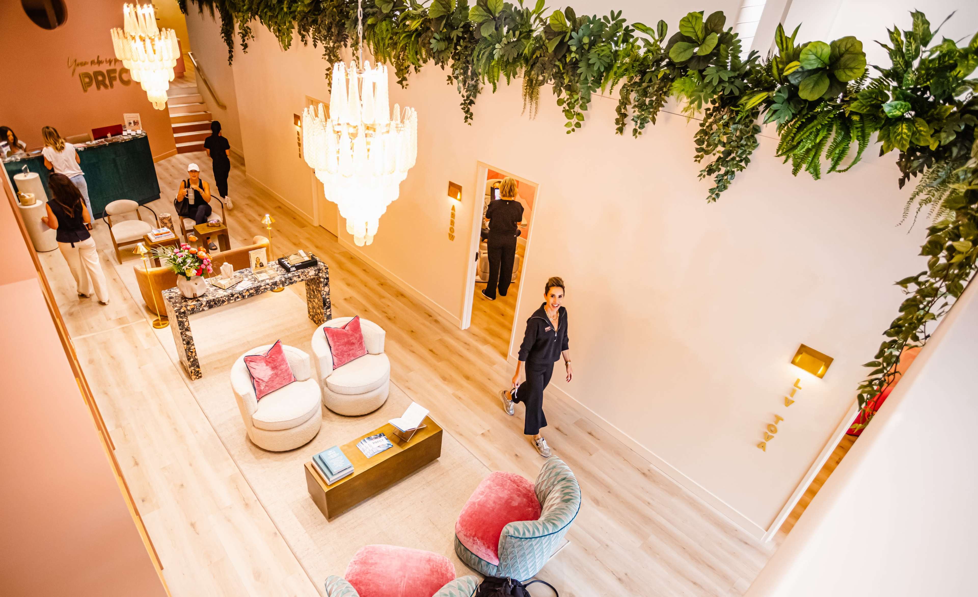 The image shows a spacious, elegantly designed interior with lush greenery adorning the ceiling, plush seating areas, and two large chandeliers illuminating the space.