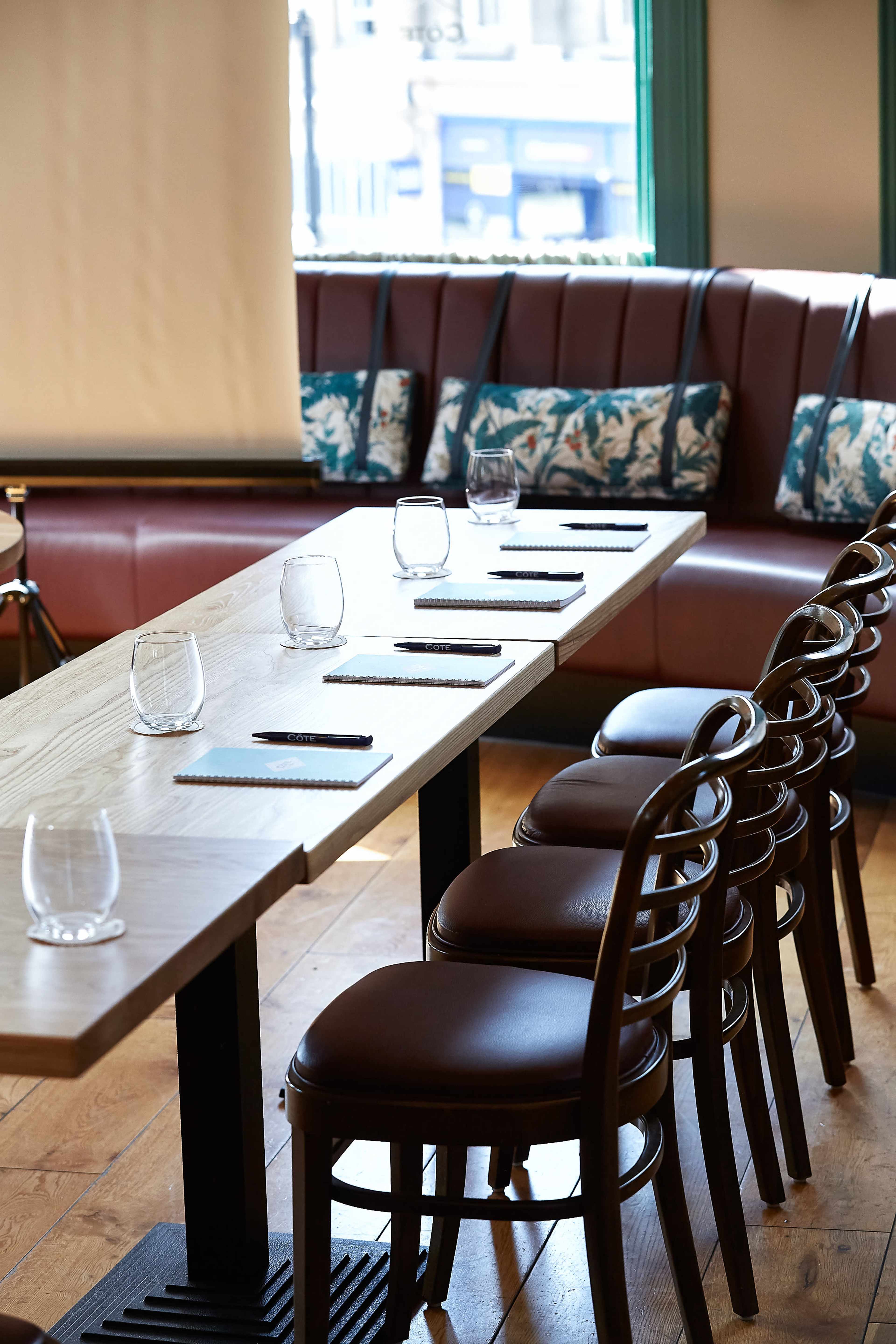 A long wooden table with place settings, glasses, and notepads is arranged in a brightly lit room with leather seating.