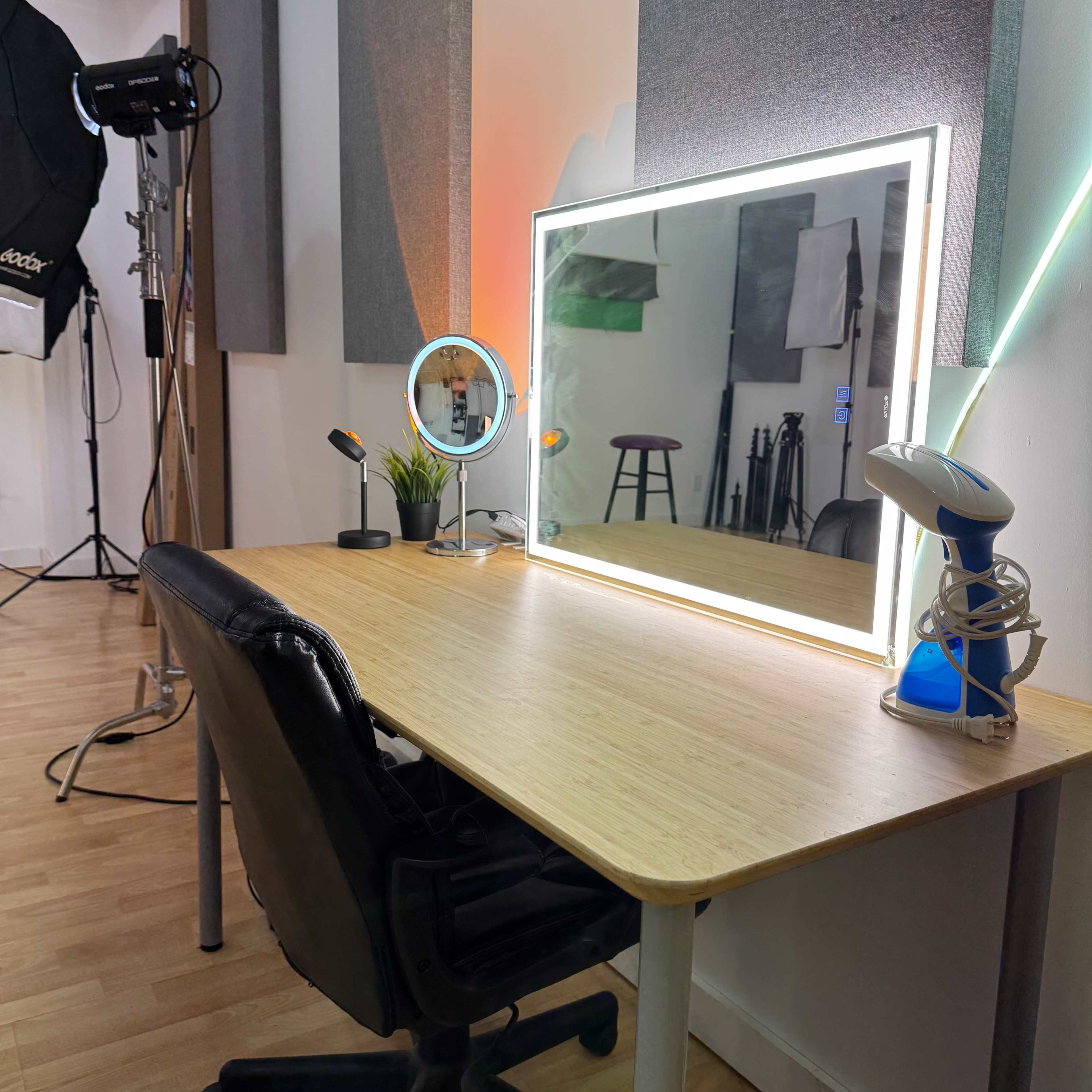 The image shows a neatly arranged workspace featuring a desk with a mirror surrounded by lights, a chair, a small plant, and various lighting equipment in the background.