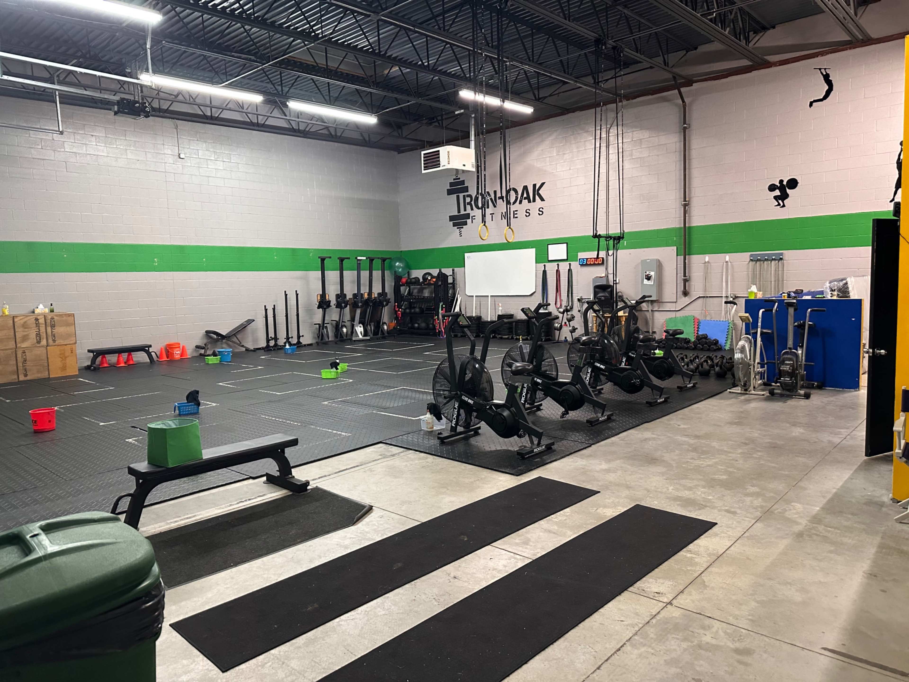 A fitness gym with rowing machines, weights, and workout equipment arranged on a rubber floor, along with storage bins and a wall featuring the "Ironoak Fitness" logo.