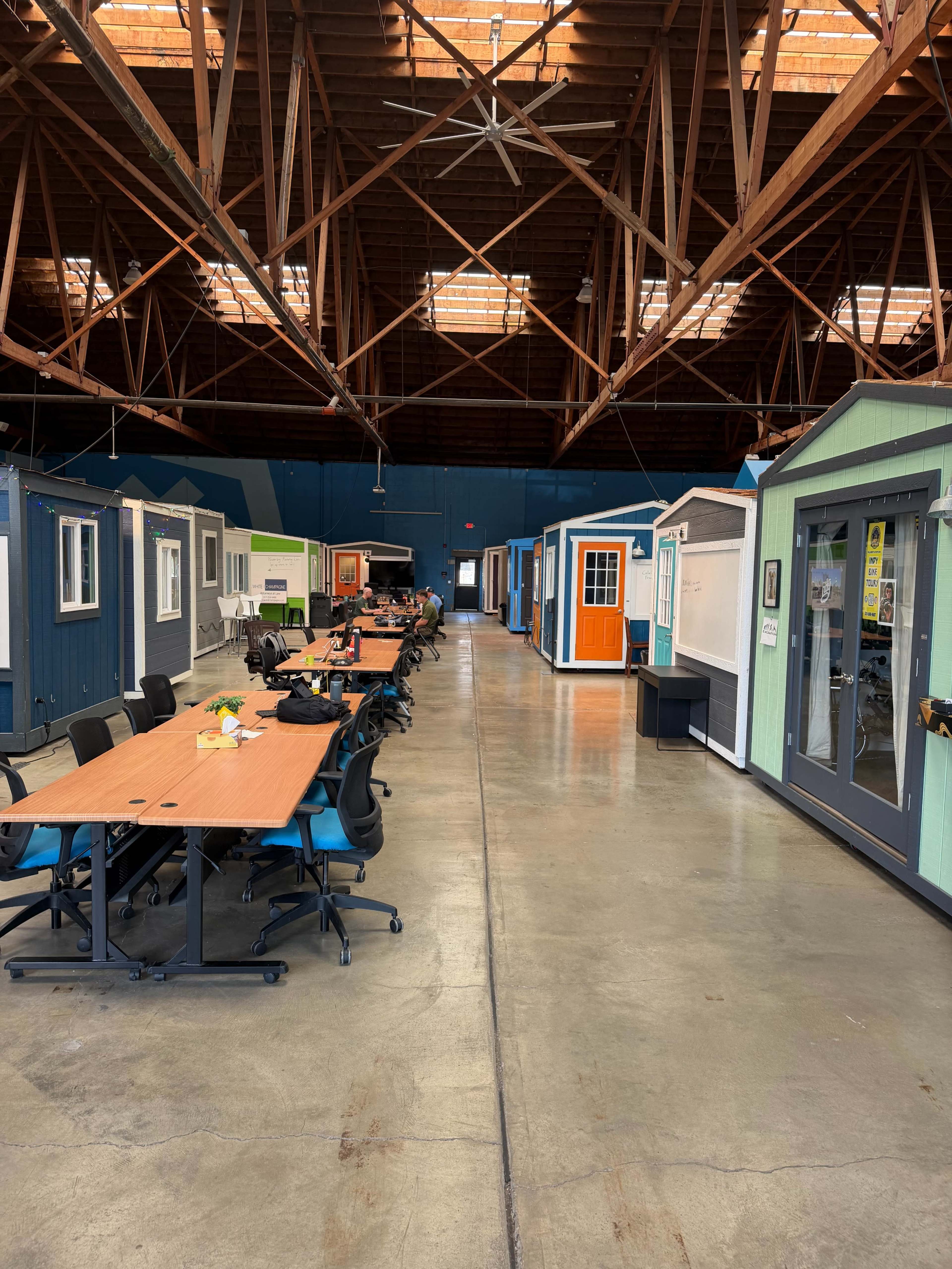 The image shows a large indoor space with several small, colorful modular structures lining the sides and a row of long tables with chairs in the center.