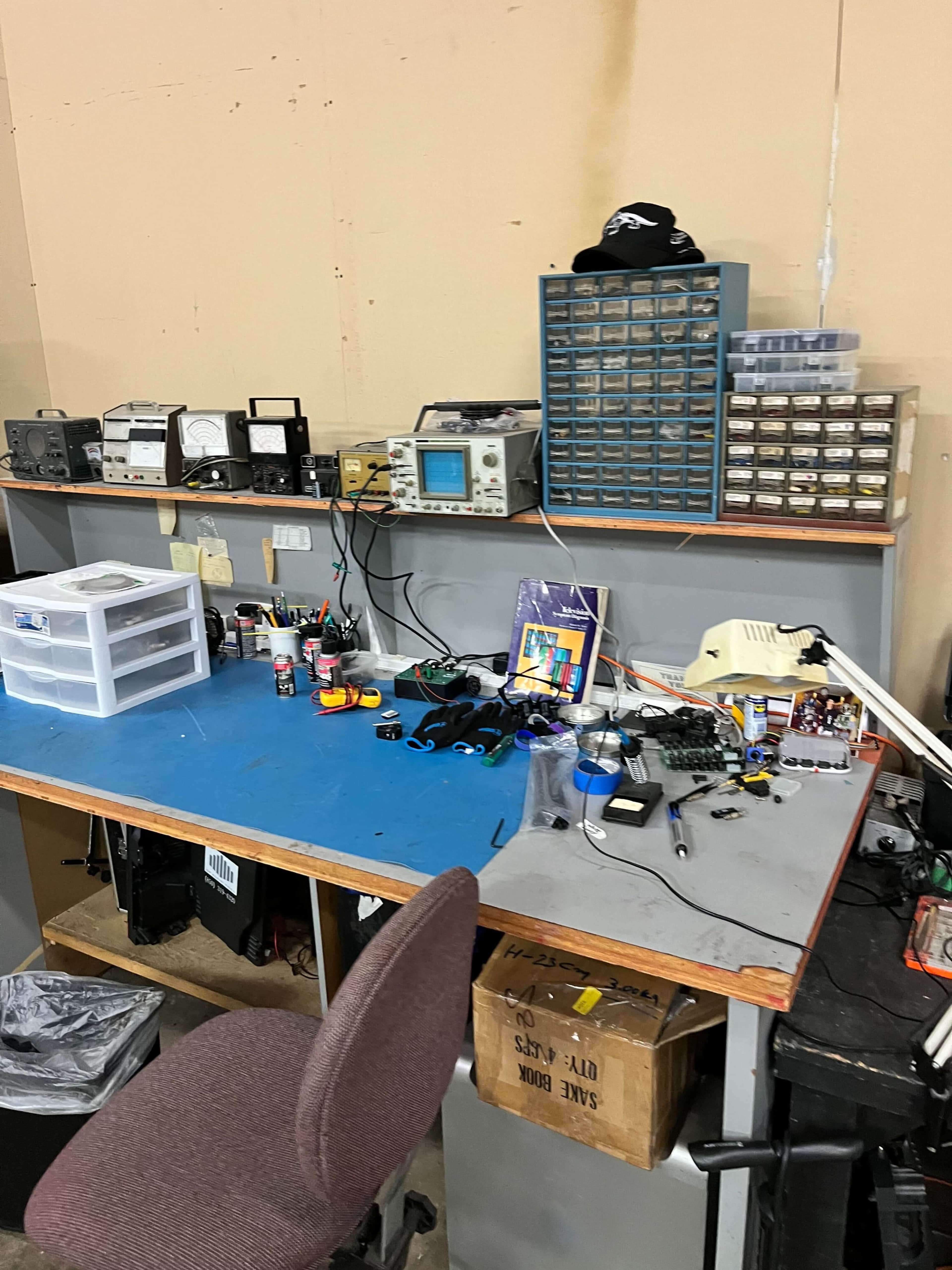 The image shows a cluttered workbench with various tools, electronic components, and storage units organized against a wall.