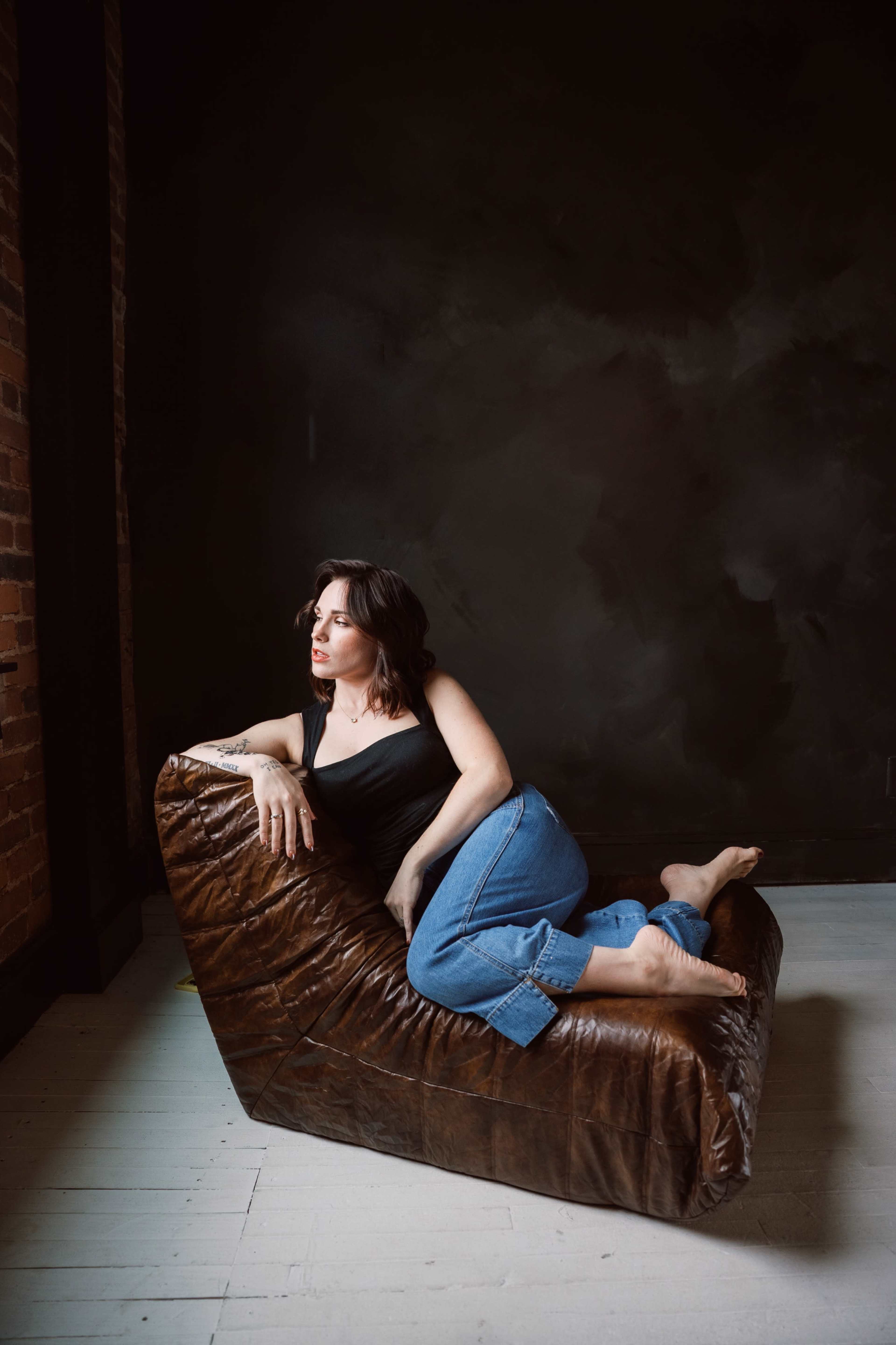 A woman reclines on a brown leather chair against a dark wall.