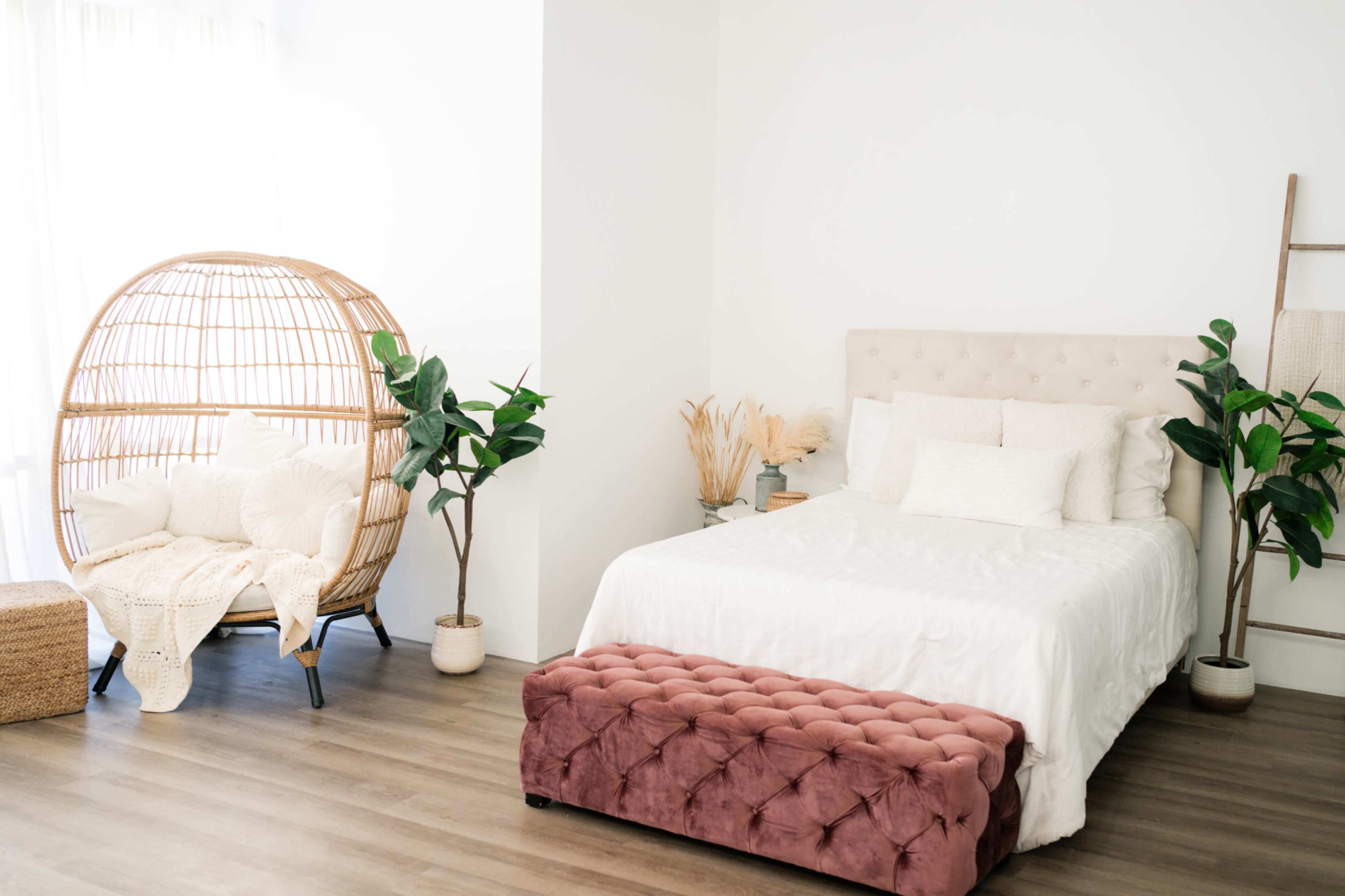 A light and airy bedroom features a large bed with white bedding, a tufted pink bench at the foot, and a woven chair surrounded by potted plants.
