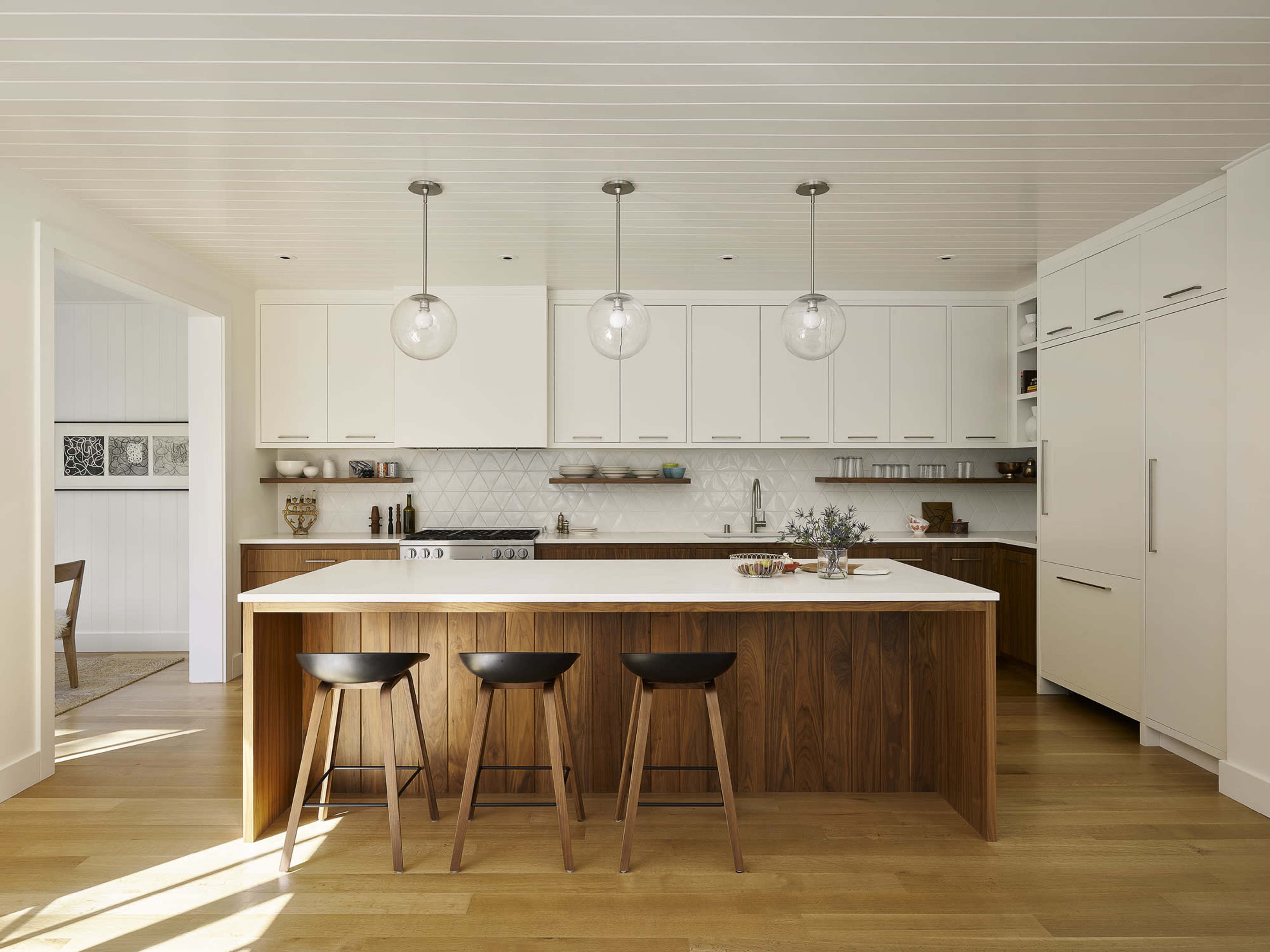 A modern kitchen features white cabinetry, a large wooden island with three black stools, and pendant lighting.