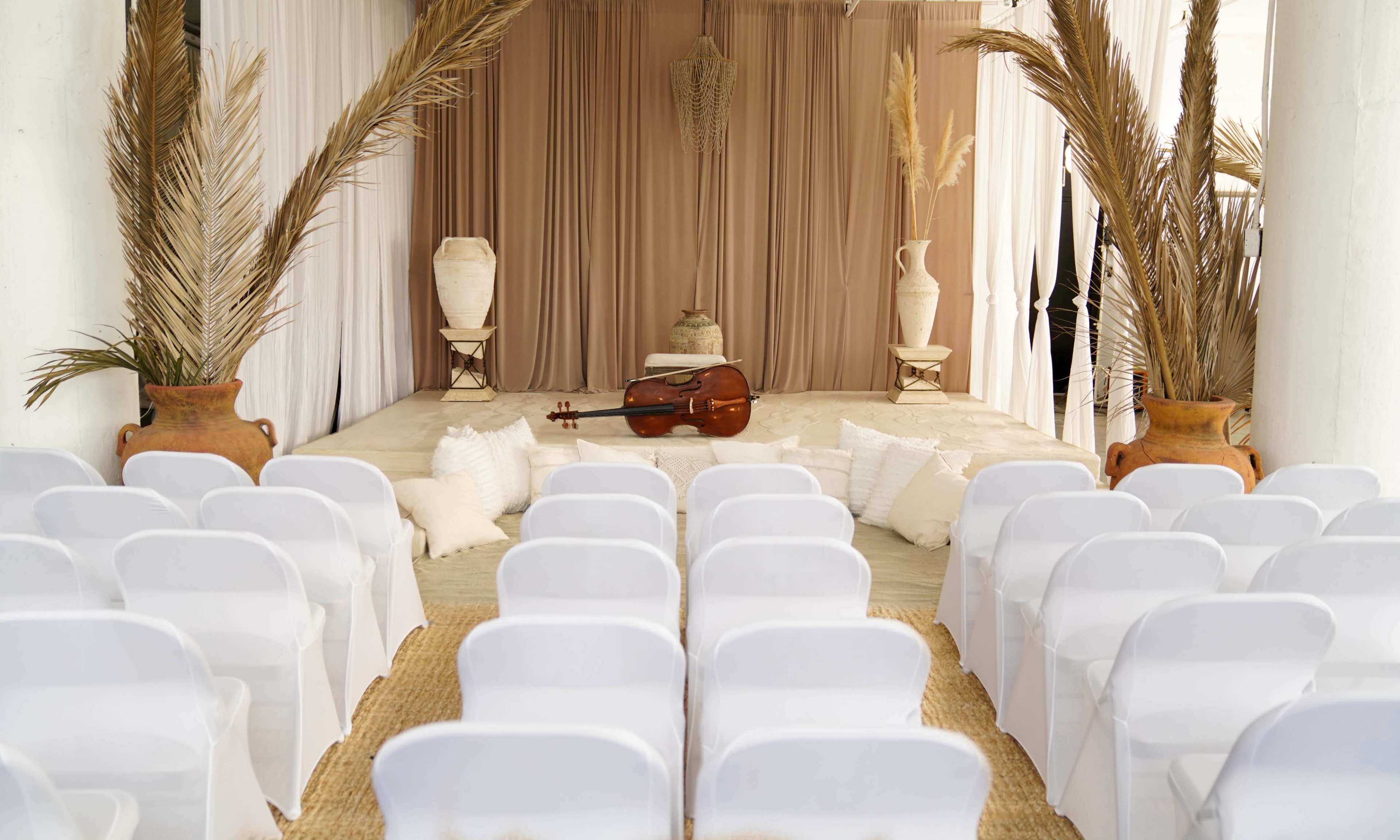 A neatly arranged indoor venue features white chairs facing a stage adorned with drapes, decorative vases, and a cello positioned centrally on a light-colored carpet.