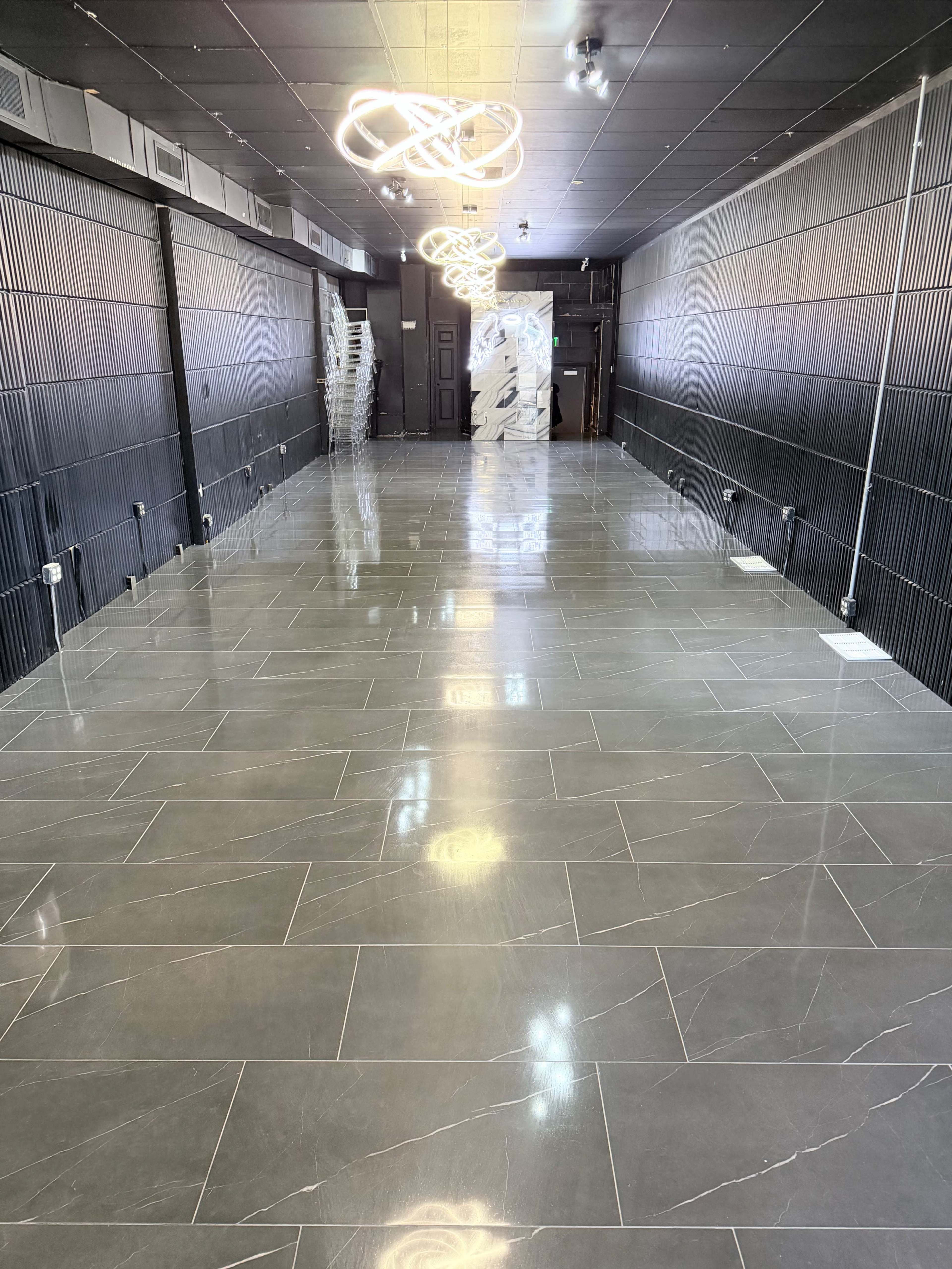 The image shows a long and spacious modern room with polished grey tile flooring and geometric light fixtures hanging from the ceiling.
