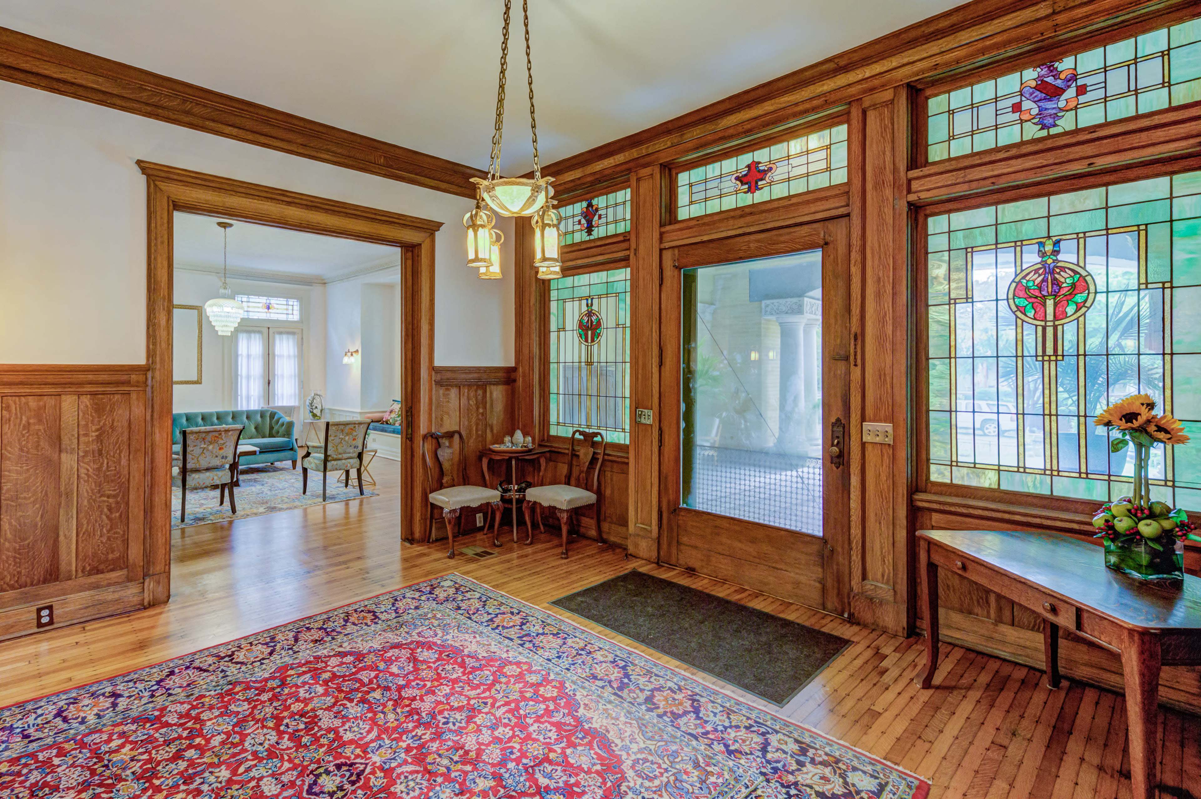 A spacious entryway with stained glass windows, wooden paneling, and a patterned area rug, leading to a brightly lit sitting area.