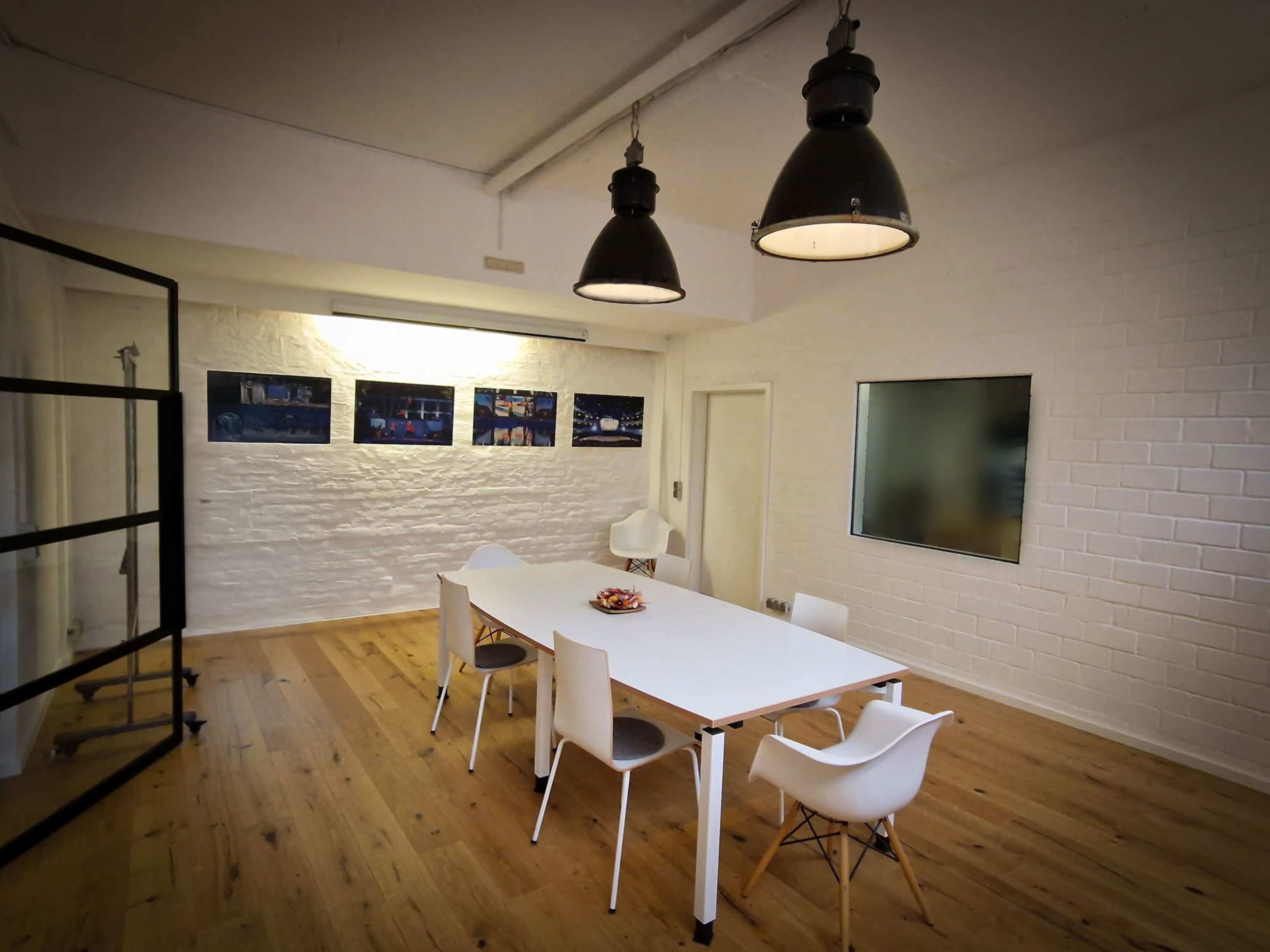 A bright meeting room features a white table surrounded by chairs, with art displayed on the walls and large pendant lights overhead.