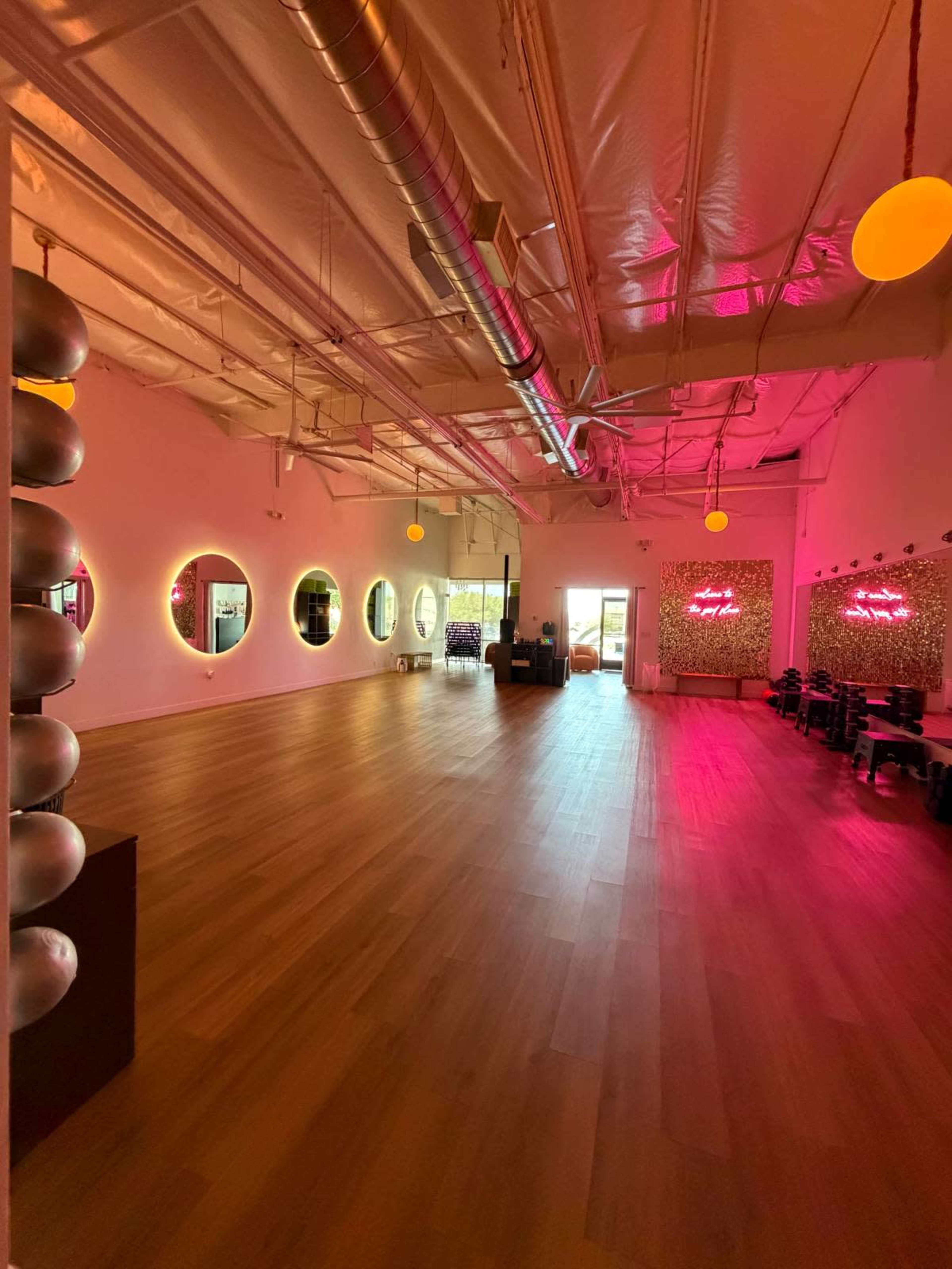 The image shows a spacious studio with wooden flooring, large circular mirrors on the walls, and ambient pink lighting.