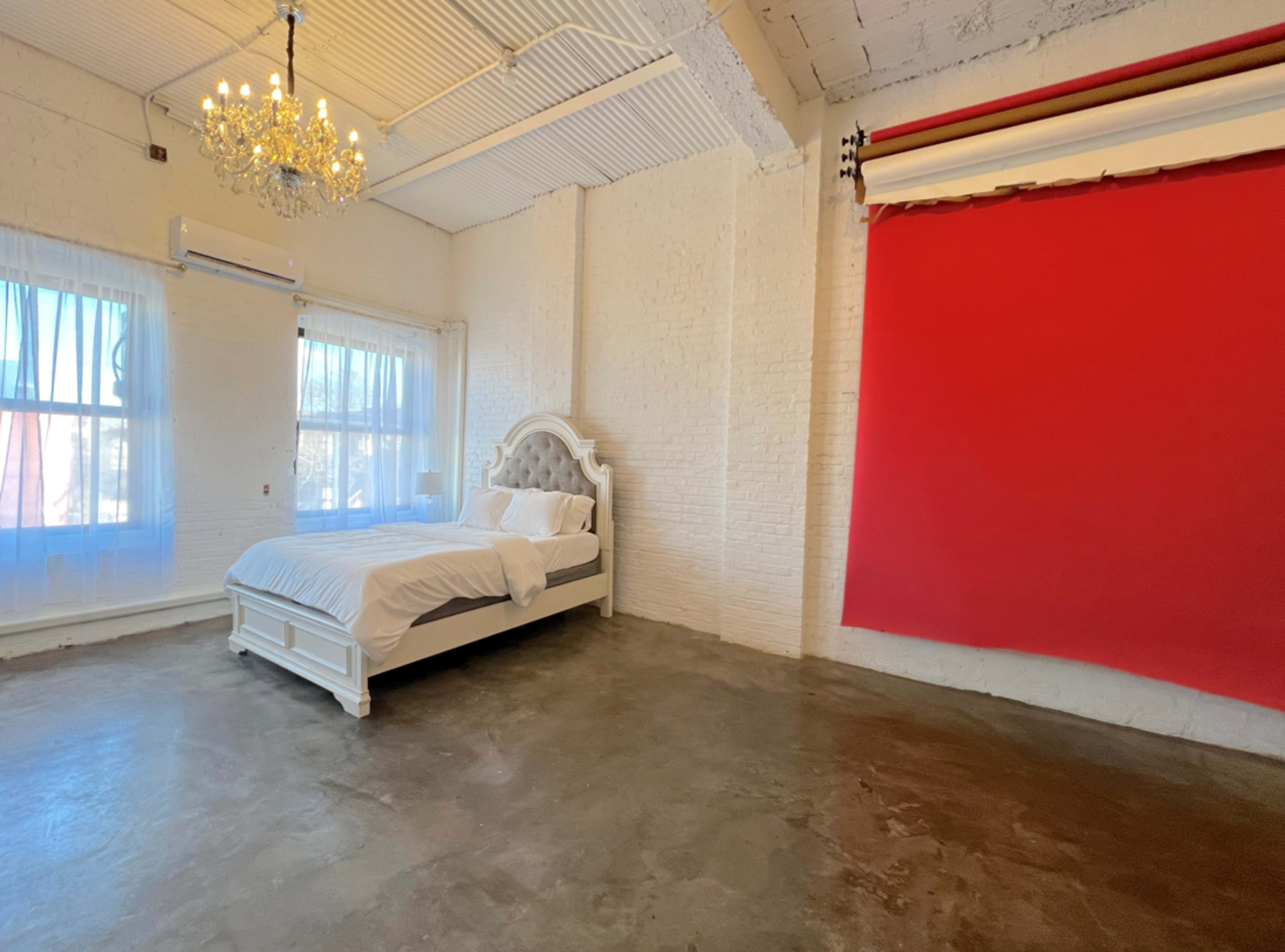 A minimalist bedroom with a white bed, large windows, and a red backdrop on one wall.