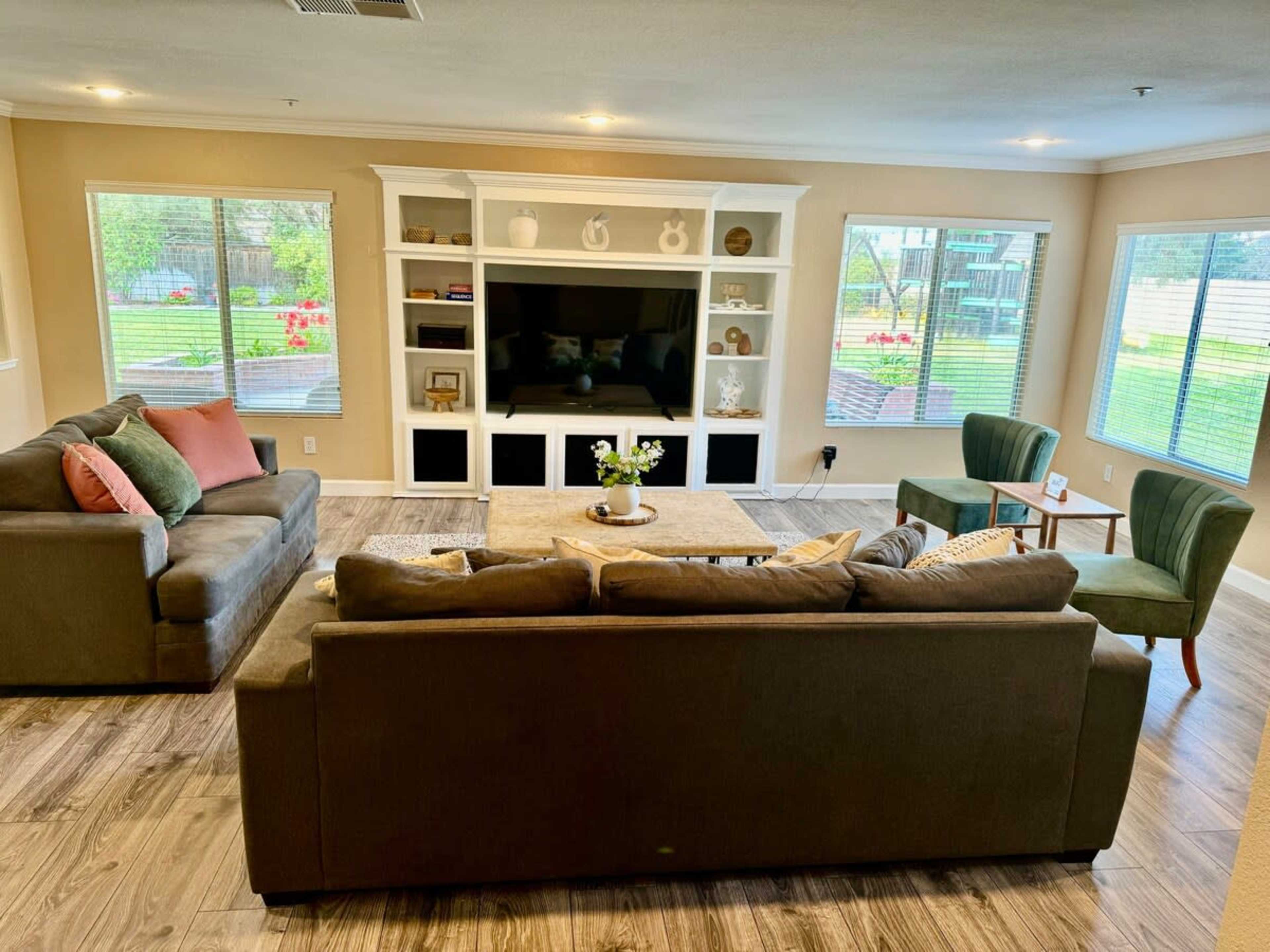 The image depicts a living room with two sofas, a coffee table, and a TV unit, surrounded by large windows that offer a view of greenery outside.