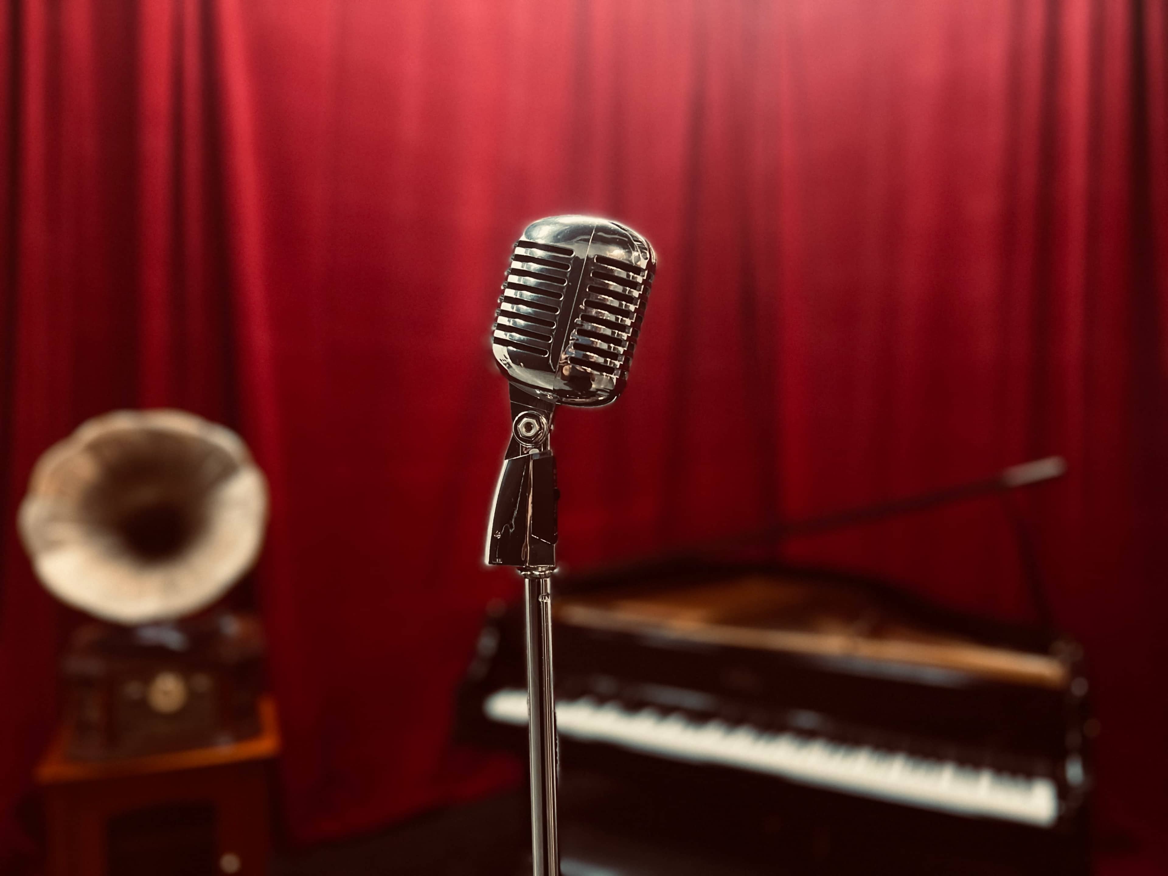 A vintage microphone stands in front of a red curtain, with a grand piano and a gramophone visible in the background.