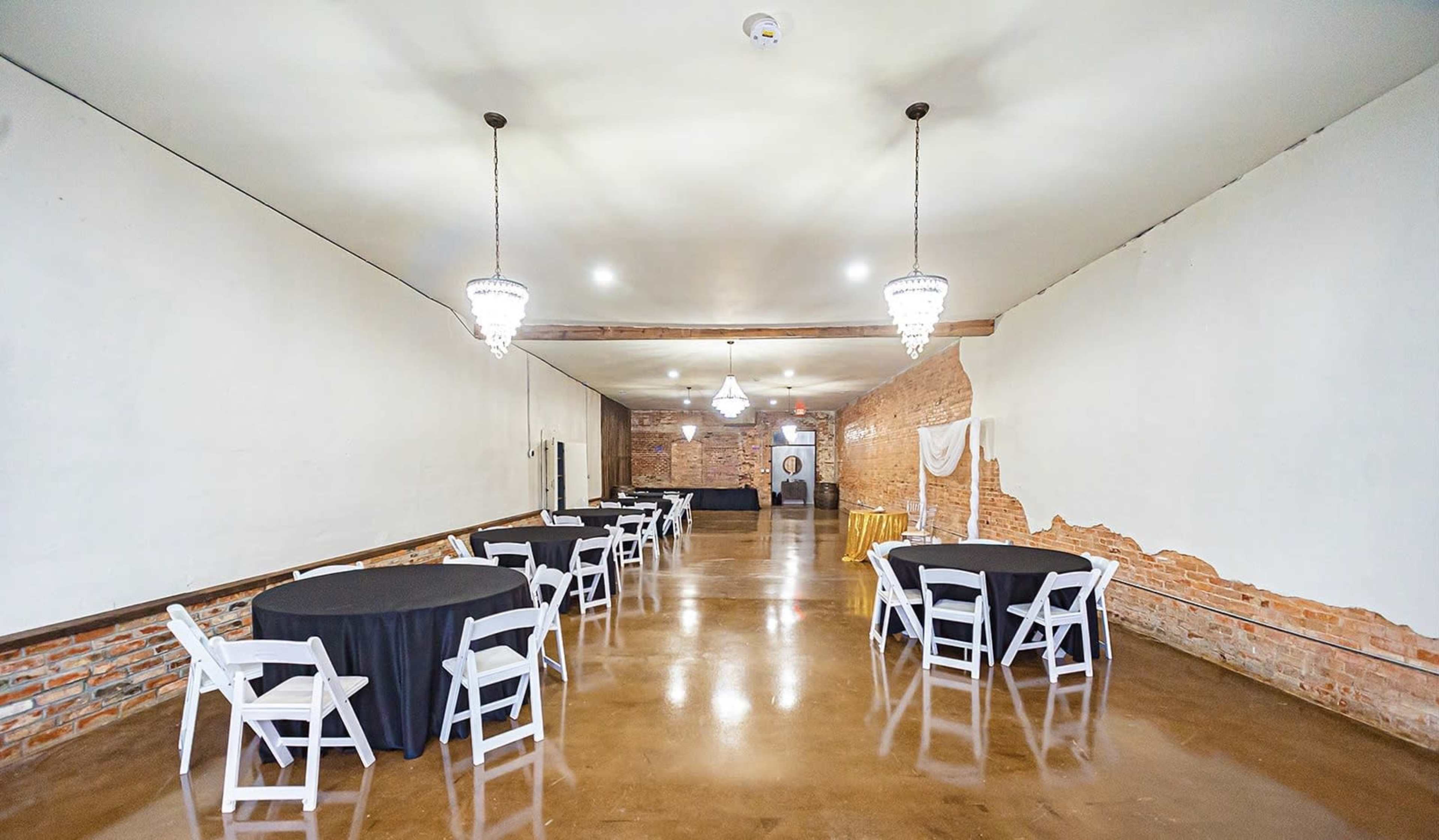 A spacious event venue with several round tables covered in black cloth and lit by chandeliers, featuring exposed brick walls and a polished concrete floor.