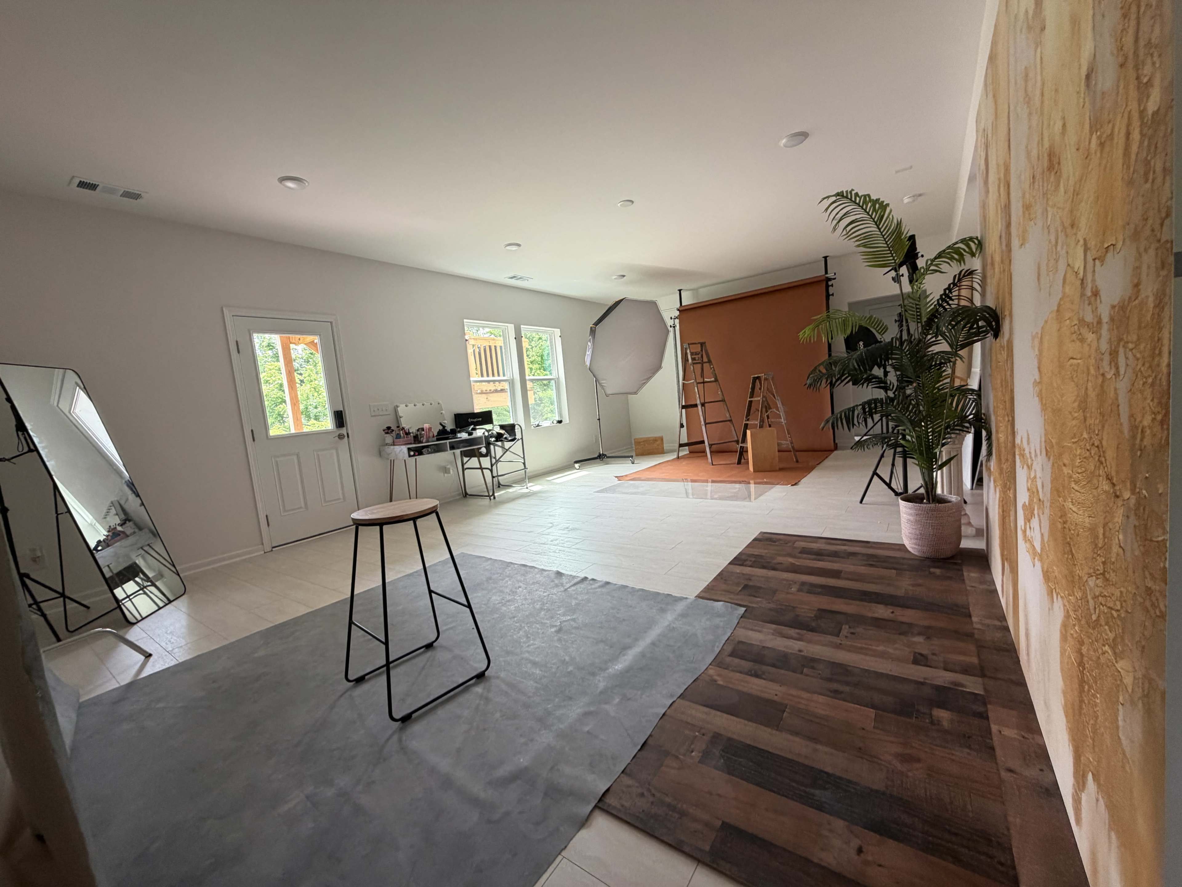 The image shows a well-lit studio space featuring a wooden floor, a gray rug, a ladder, a backdrop for photography, and a large mirror.