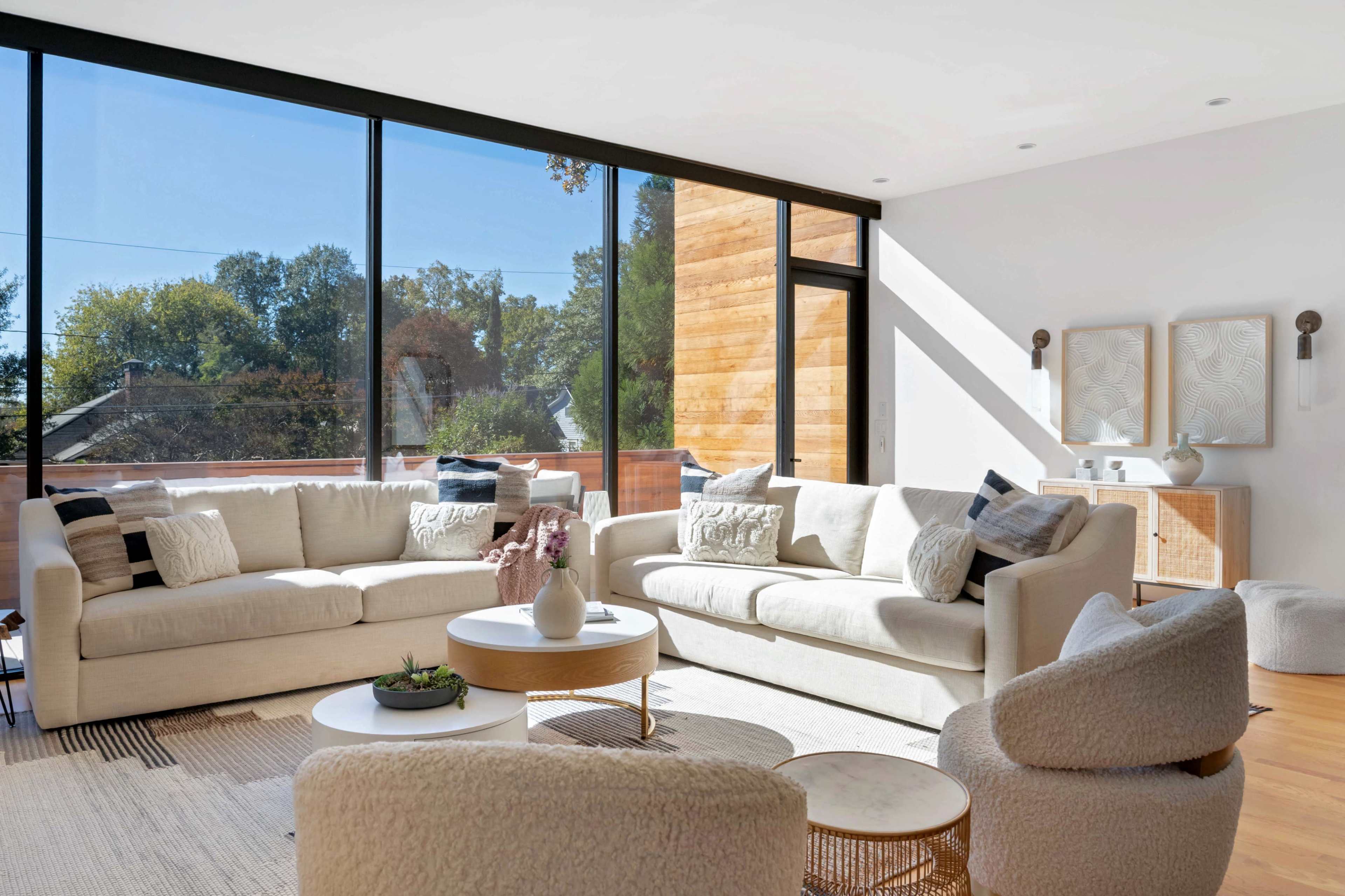 The image features a modern living room with large windows allowing natural light, two beige sofas, a round coffee table, and decorative items placed around the space.
