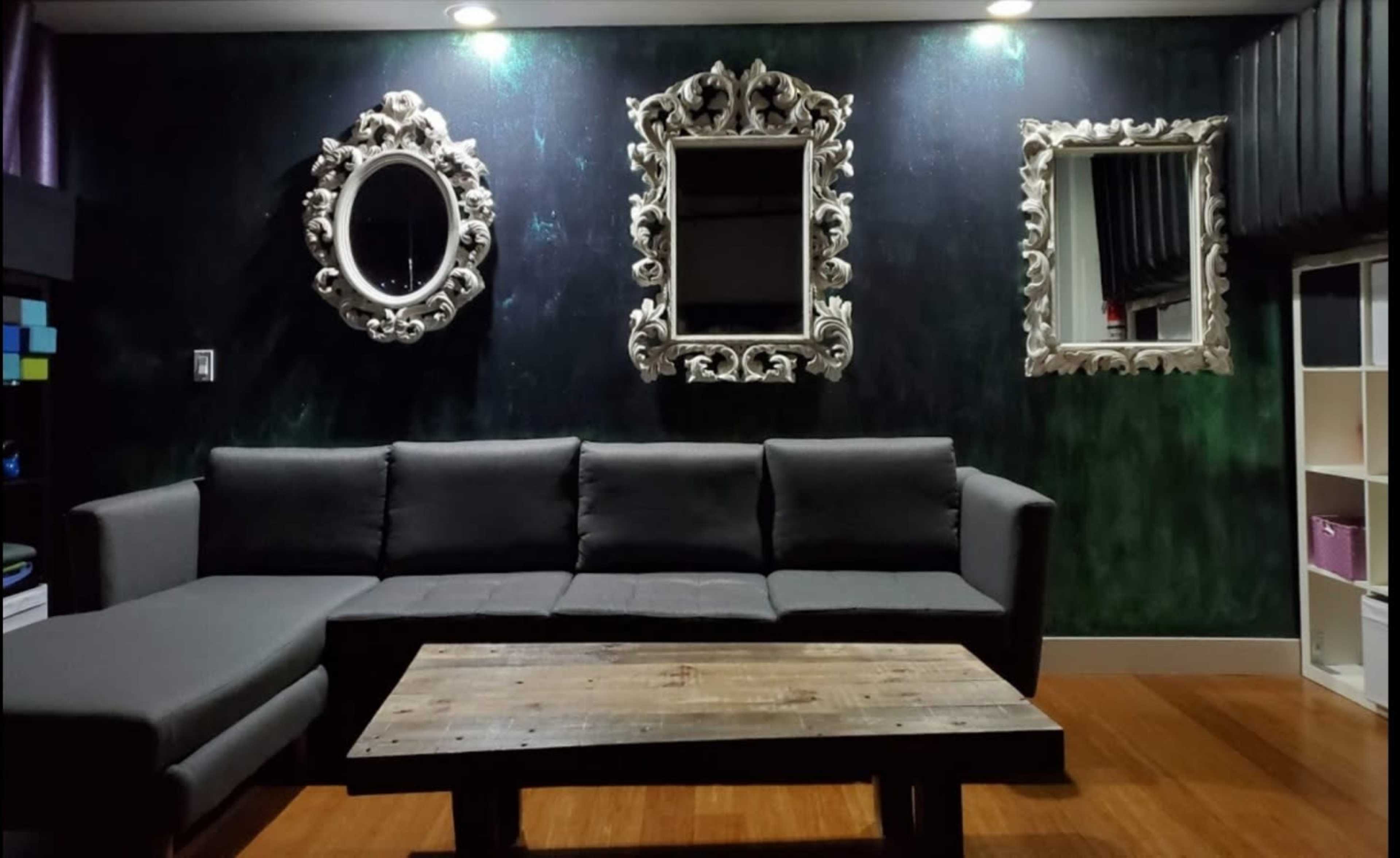 A modern living room with a dark green wall, an L-shaped gray sofa, a wooden coffee table, and three ornate mirrors mounted on the wall.